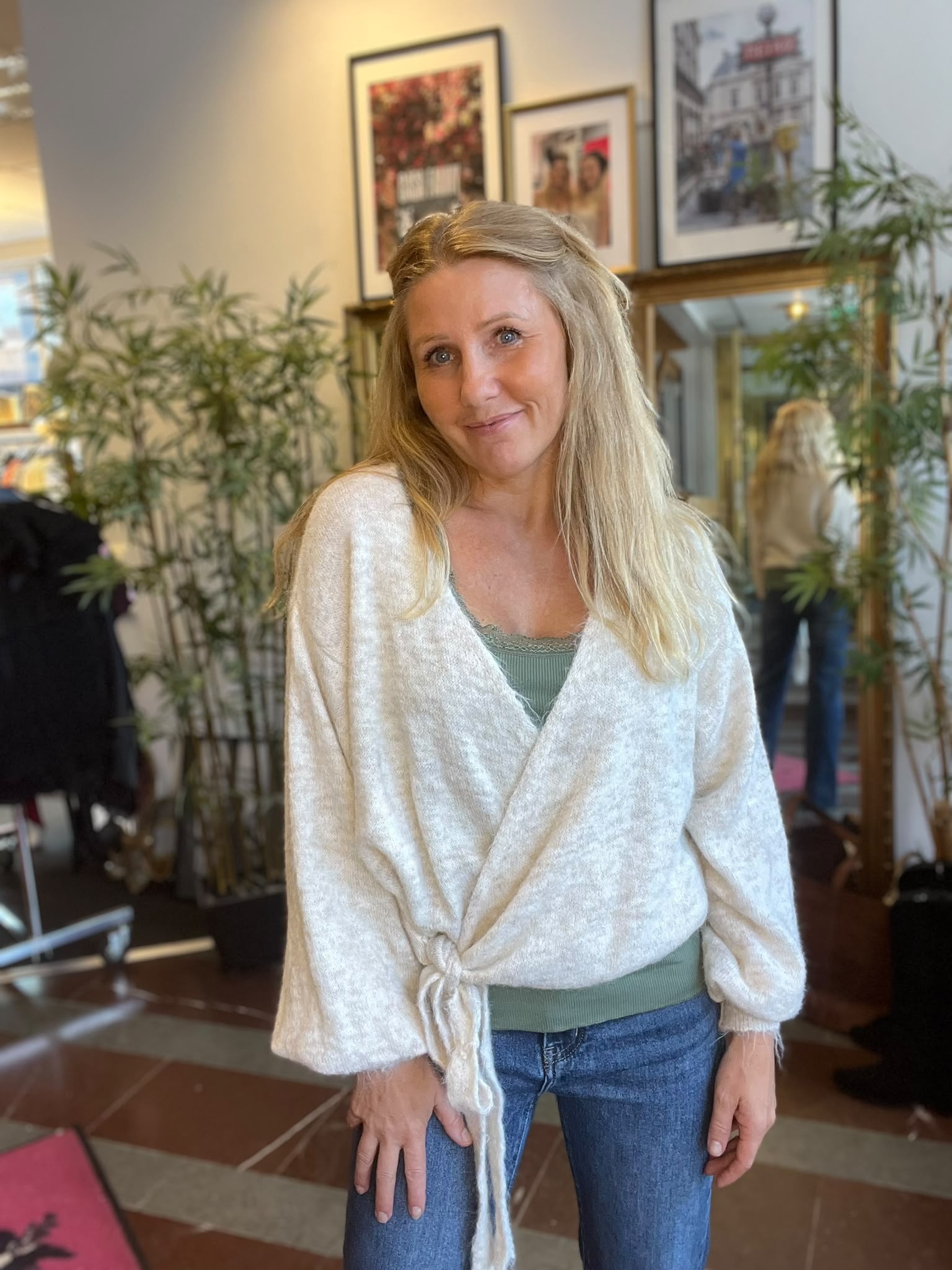 Leende kvinna med långt blont hår står vänd mot kameran och lutar sig lätt framåt. Hon bär en vit omlottkofta över grön topp och jeans, i en inomhusmiljö med växter, spegel och inramade tavlor.
