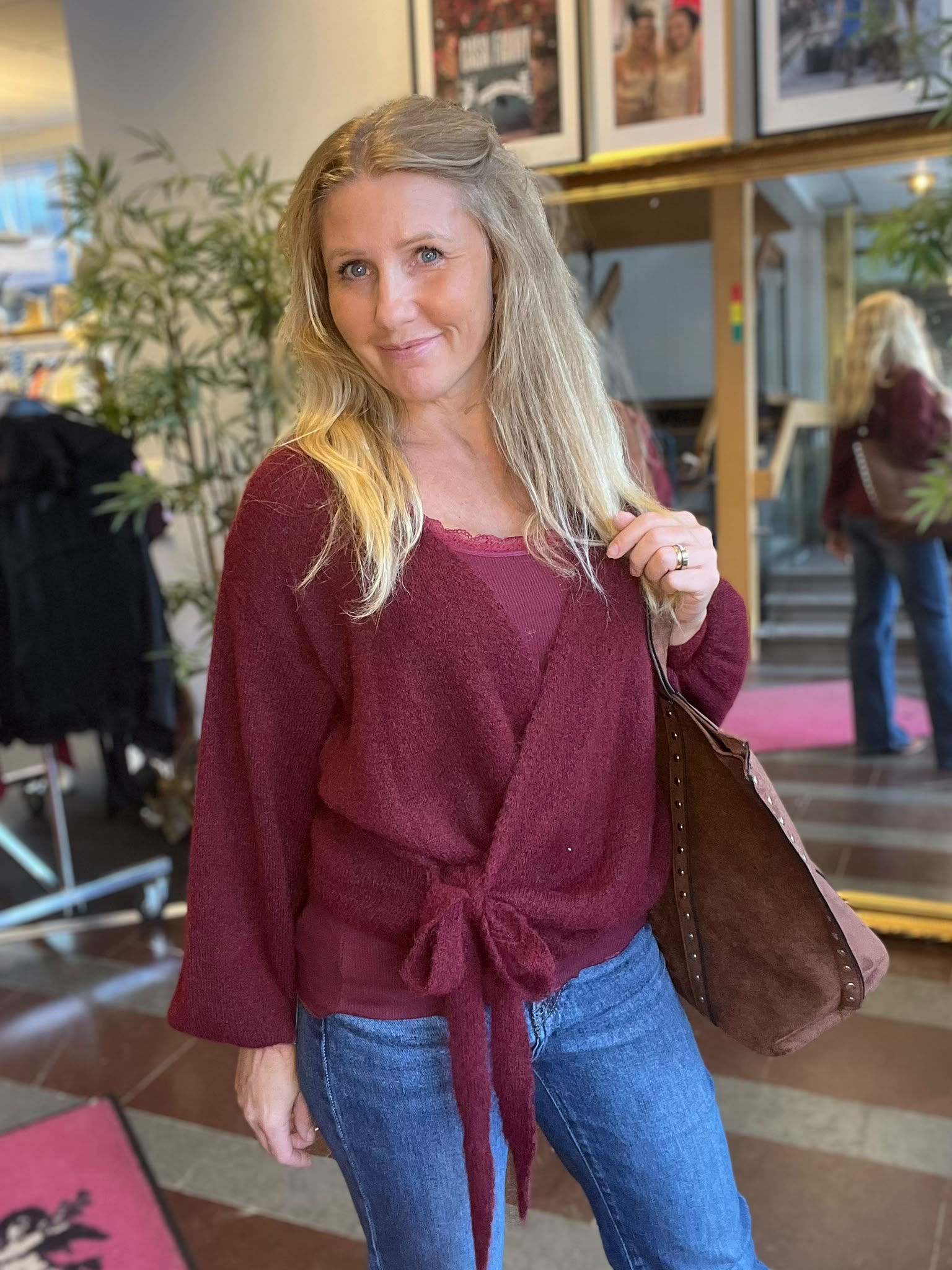 Leende blond kvinna poserar inomhus och håller en brun axelväska, klädd i vinröd knuten topp och blå jeans. I bakgrunden syns växter, inramade foton och en spegel som reflekterar rummet.