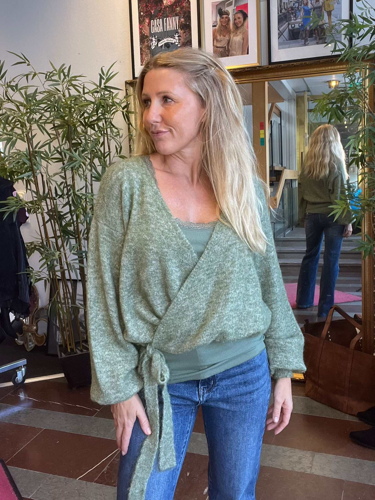 Blond kvinna i grön omlottkofta och jeans står inomhus och tittar åt sidan. Bakom henne syns en stor guldramad spegel med hennes rygg, gröna växter och inramade foton; texten “CASA FANNY”.
