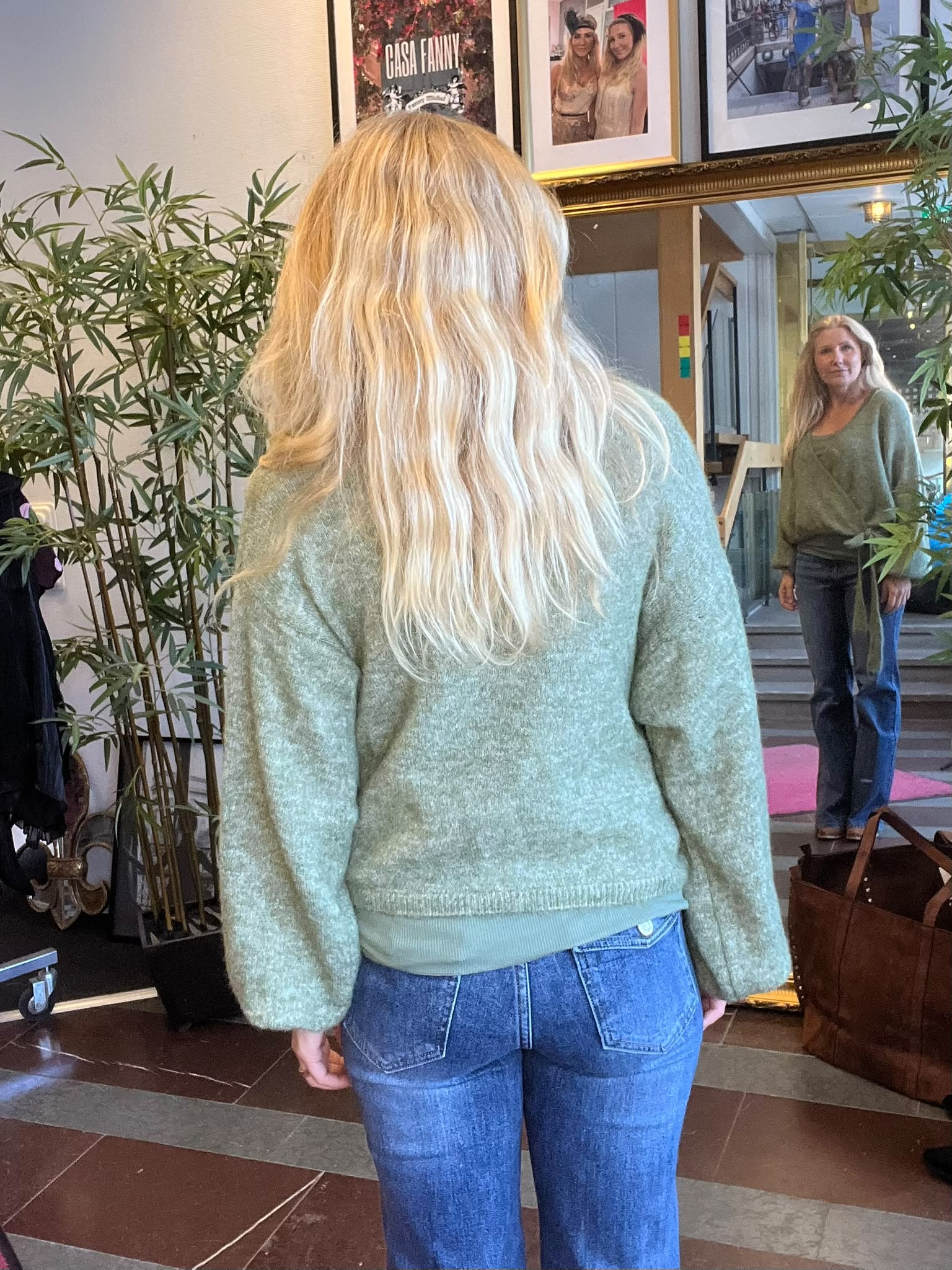 Kvinna med långt blont hår står med ryggen mot kameran och tittar på sin spegelbild i ett stort guldram. Hon bär grön stickad tröja och jeans; växter och tavlor syns, inklusive texten ”CASA FANNY”.