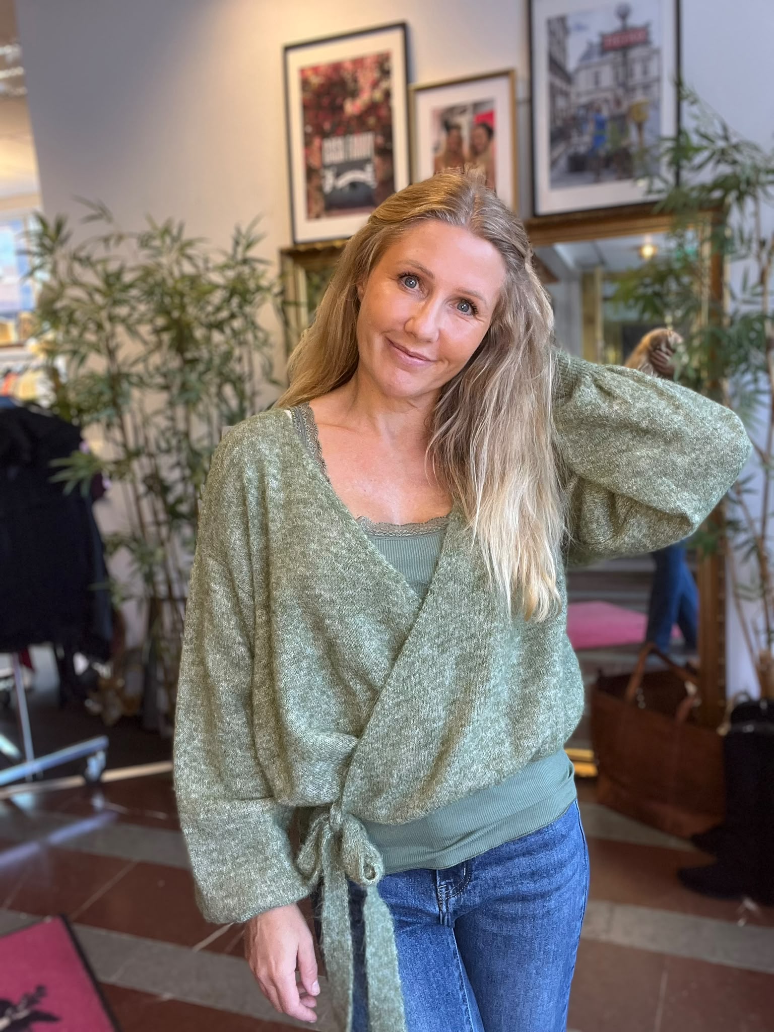 Leende kvinna med långt blont hår står avslappnat och håller ena handen bakom huvudet, klädd i grön omlottkofta och jeans, i ett café- eller vardagsrumsliknande rum med växter och inramade foton.