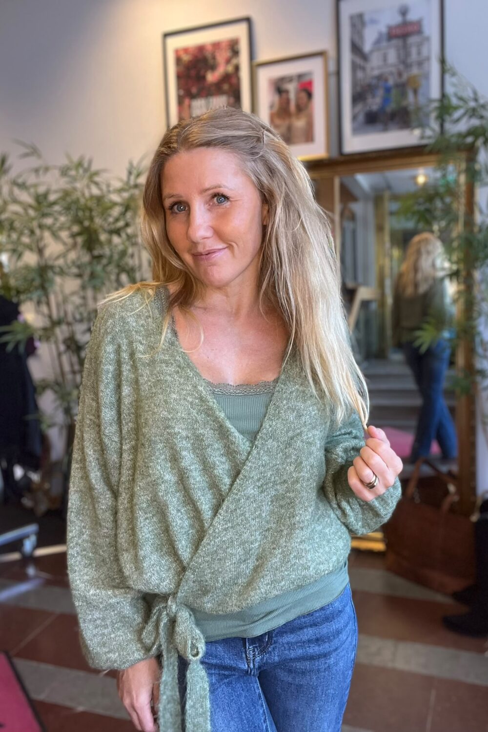Leende kvinna med långt blont hår står inomhus och håller i sin gröna omlottkofta över en grön topp och jeans. I bakgrunden syns krukväxter, inramade tavlor, en spegel och en soffa.