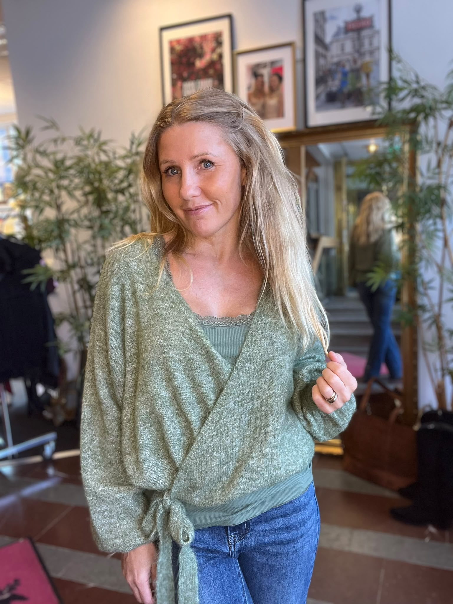 Leende kvinna med långt blont hår står inomhus och håller i sin gröna omlottkofta över en grön topp och jeans. I bakgrunden syns krukväxter, inramade tavlor, en spegel och en soffa.