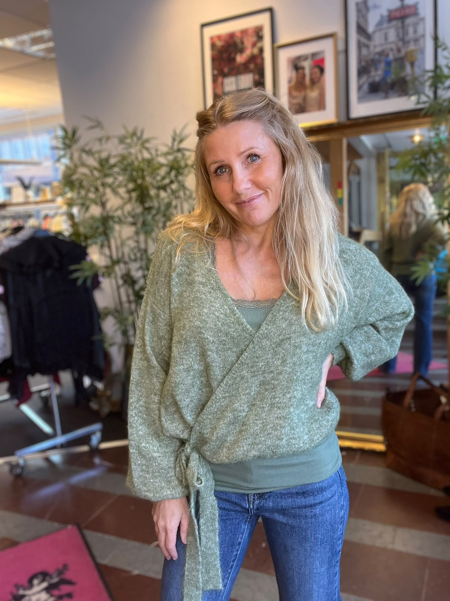 Leende blond kvinna står lätt lutad med handen på höften och ser in i kameran. Hon bär grön omlottkofta och jeans. Bakom syns en inomhusmiljö med spegel, växter och inramade foton på väggen.