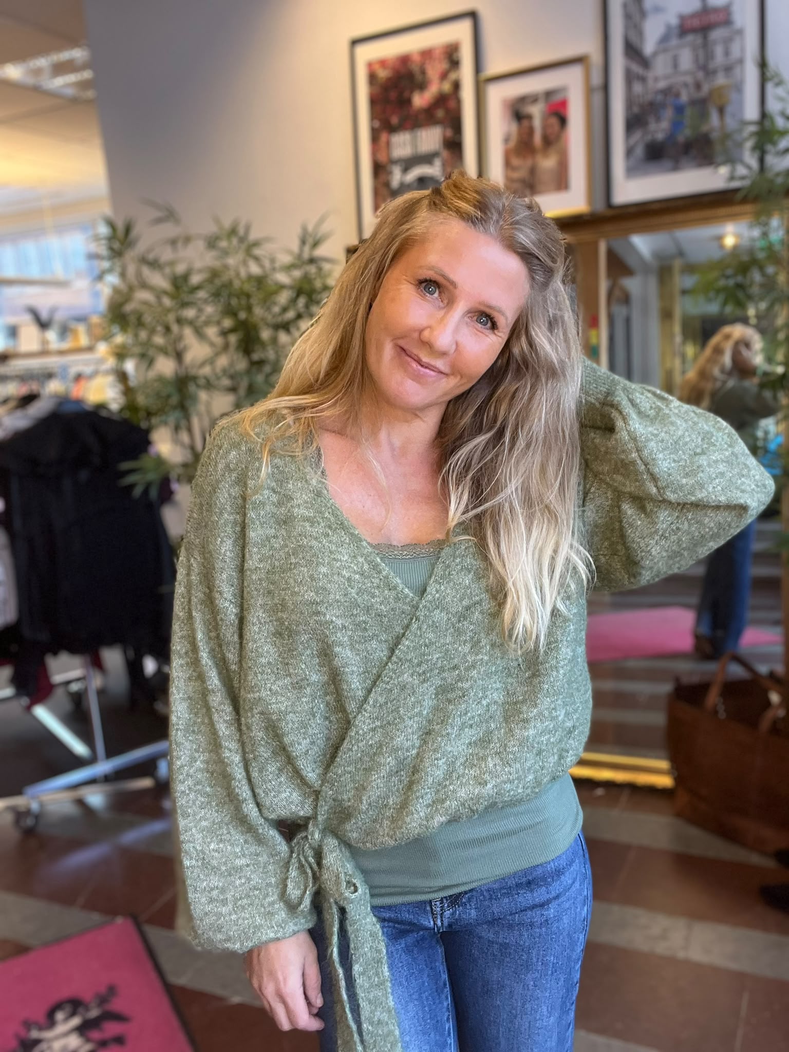 Leende kvinna med långt blont hår lutar huvudet och håller ena handen bakom nacken, klädd i grön stickad tröja och jeans. Hon står i ett ljust rum med växter och inramade fotografier på väggen.