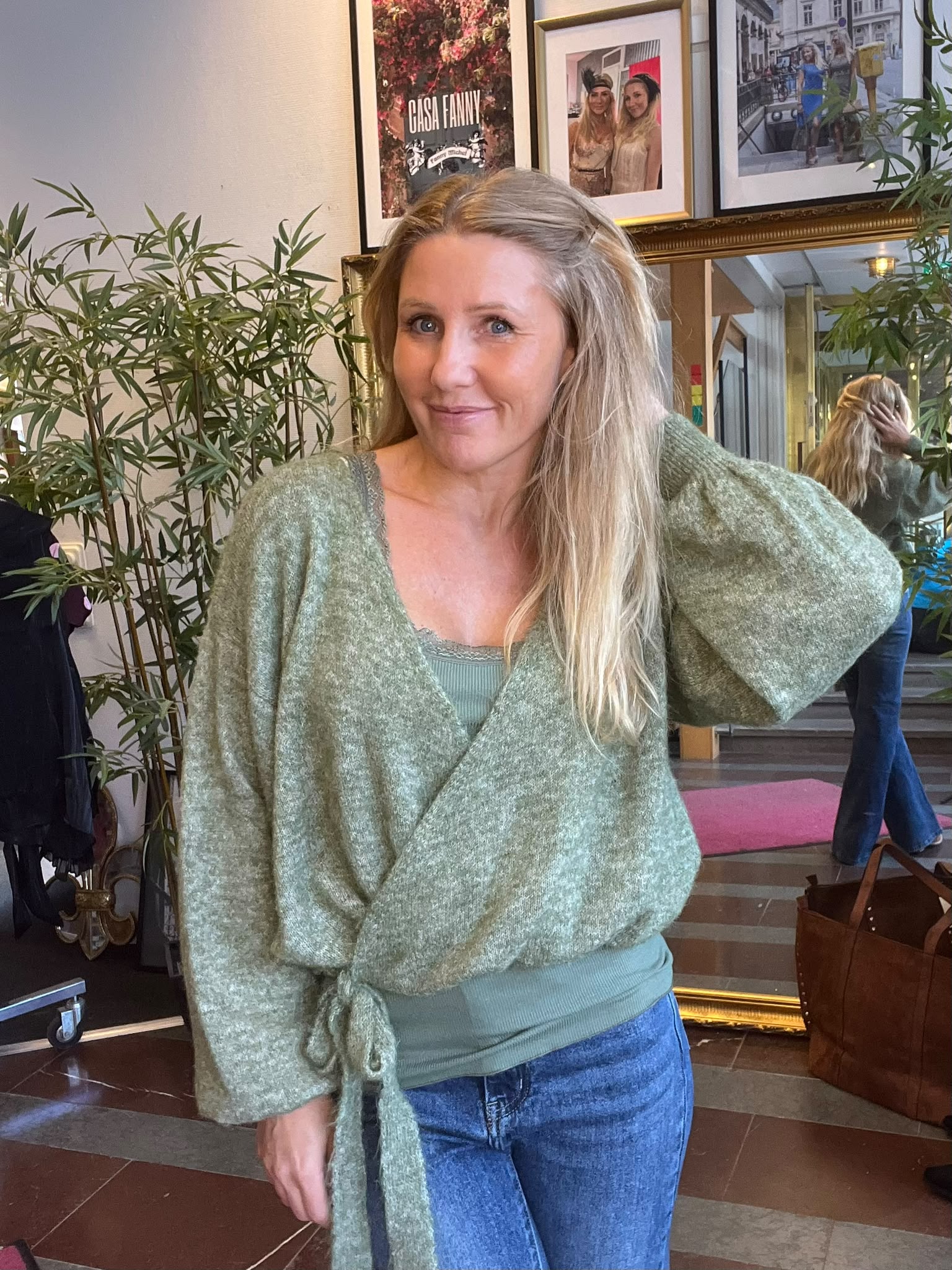 Leende kvinna i grön omlottkofta står inomhus och håller ena handen i håret. Bakom syns gröna växter, en spegel och inramade foton; en affisch med texten “CASA FANNY”.