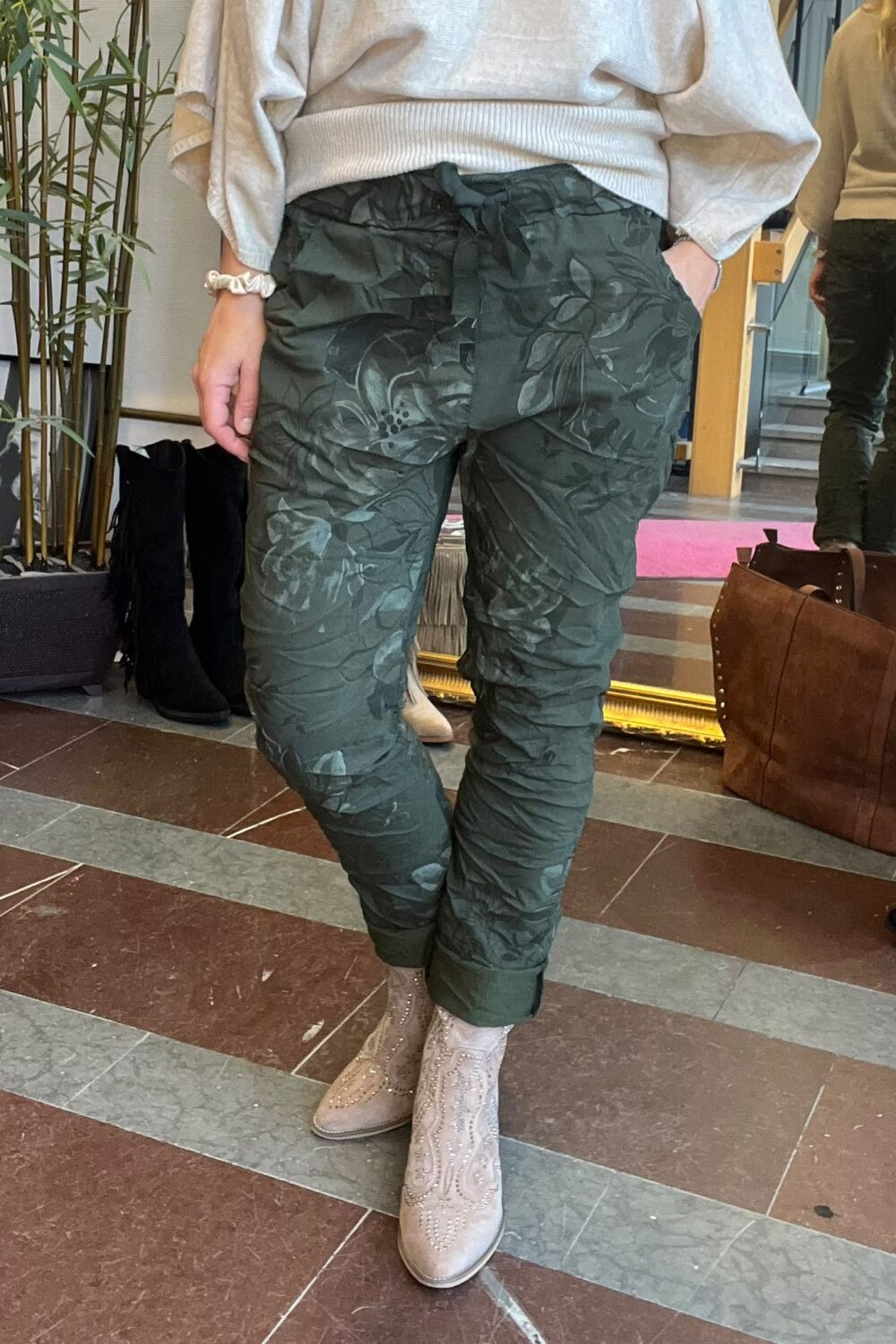 Person står med händerna i fickorna och visar mörkgröna byxor med blommigt mönster och knytband i midjan. Hen bär ljus tröja och beige, dekorerade boots i en inomhusmiljö med spegel och växter.