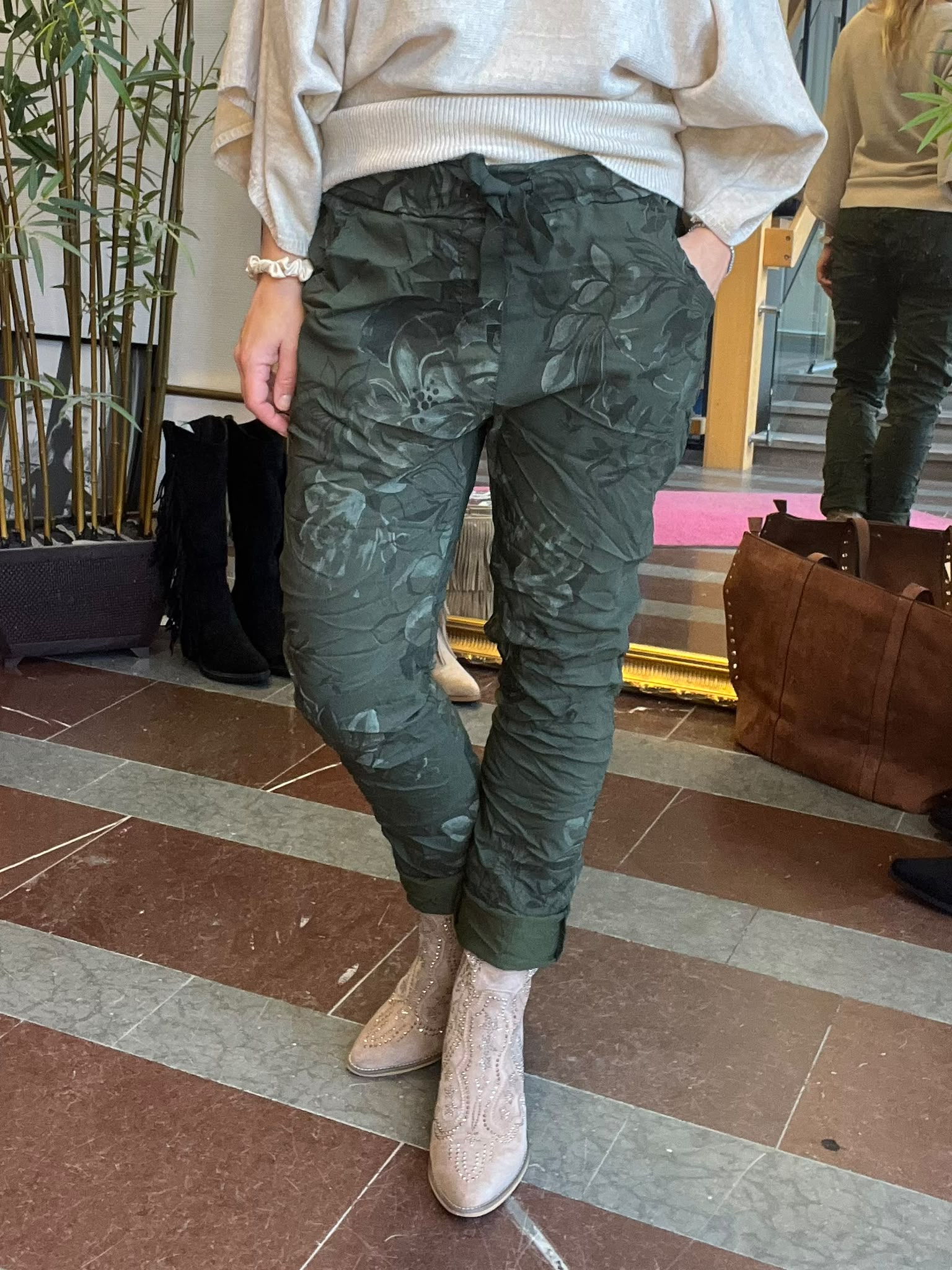 Person står med händerna i fickorna och visar mörkgröna byxor med blommigt mönster och knytband i midjan. Hen bär ljus tröja och beige, dekorerade boots i en inomhusmiljö med spegel och växter.