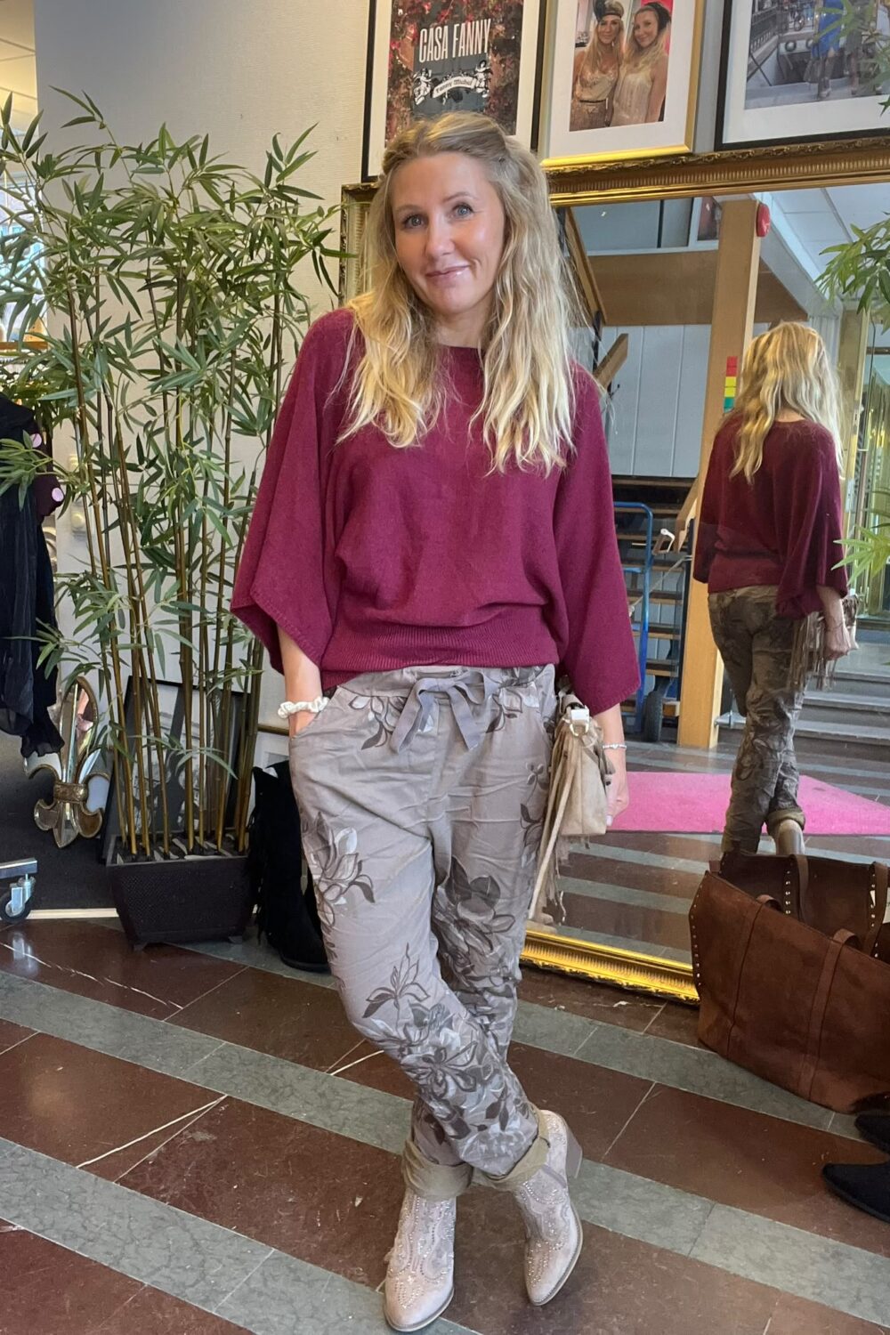 Blond kvinna står och ler med händerna i fickorna, klädd i vinröd tröja, mönstrade byxor och ljusa boots, i en inomhusmiljö med spegel och växter. Text på tavla: “CASA FANNY”.