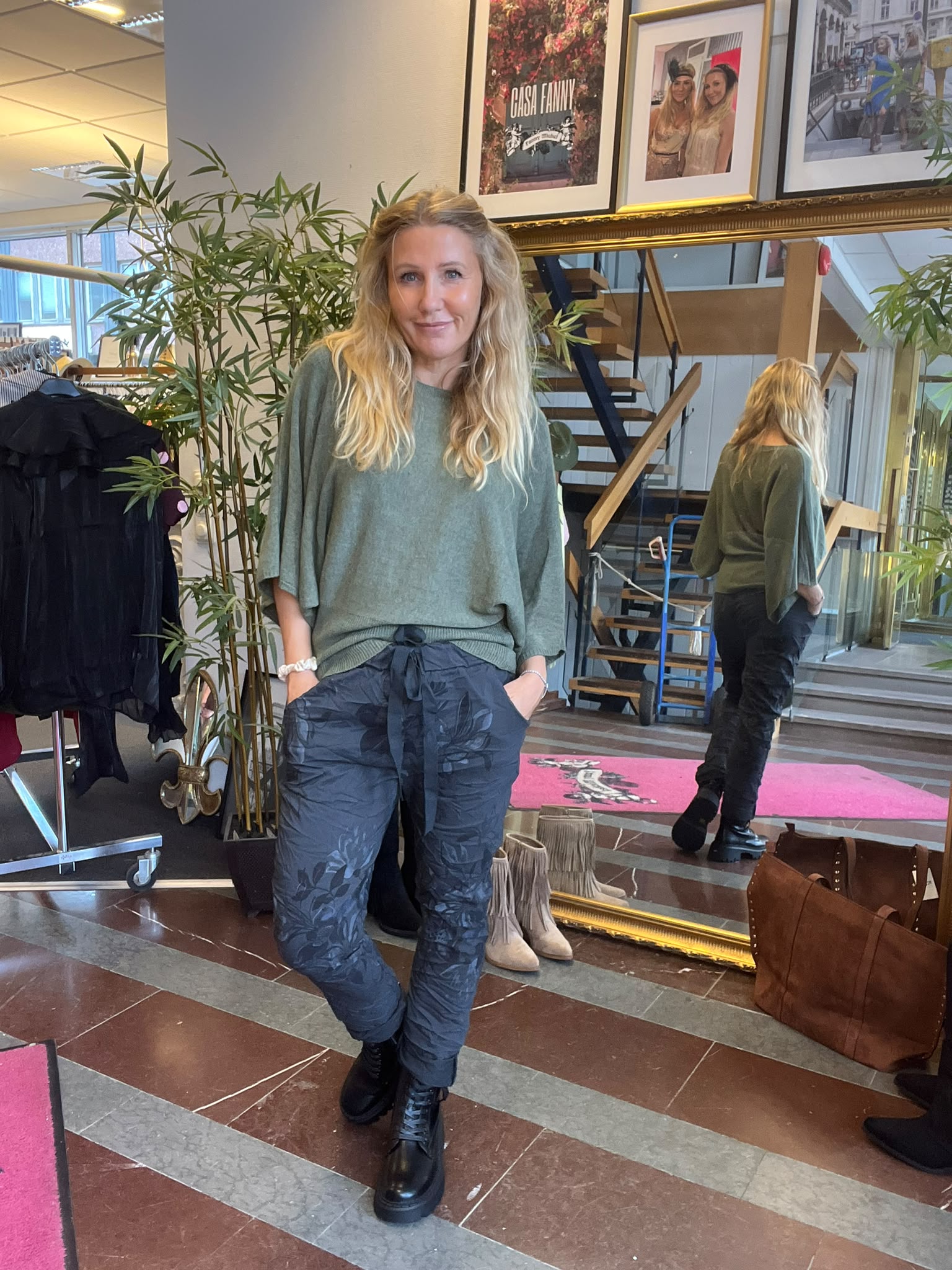 Blond kvinna står och ler med händerna i fickorna, klädd i grön topp och mönstrade byxor. Hon poserar i en butiksmiljö med stor spegel, trappa, växter och tavlor; text: ”CASA FANNY”.