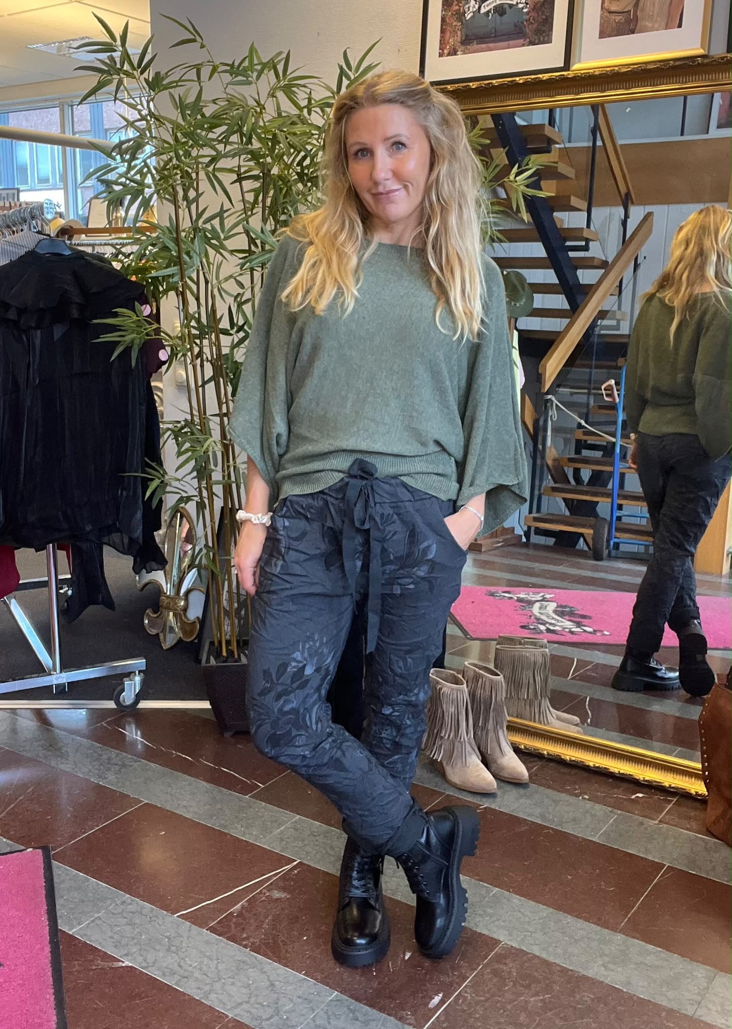 Blond kvinna står med handen i fickan och ena benet korsat, klädd i grön stickad tröja, mörka mönstrade byxor och svarta kängor. Inomhus i butiksmiljö med klädställning, stor spegel, trappa och skor.