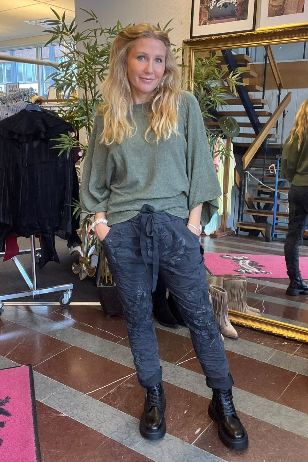 Blond kvinna poserar med händerna i fickorna och ler mot kameran, klädd i grön stickad tröja, mörka mönstrade byxor och svarta kängor. Hon står i en butik med klädställning, spegel och trappa.