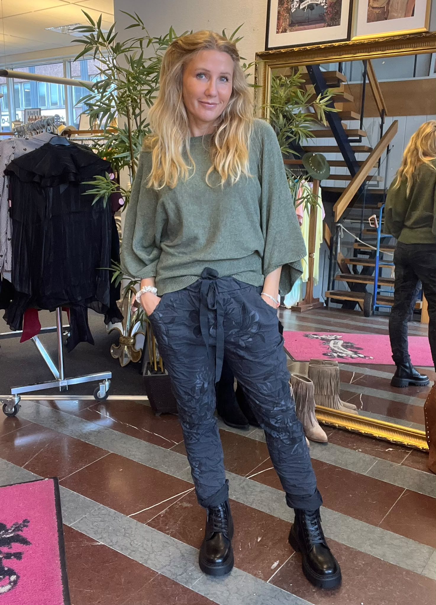 Blond kvinna poserar med händerna i fickorna och ler mot kameran, klädd i grön stickad tröja, mörka mönstrade byxor och svarta kängor. Hon står i en butik med klädställning, spegel och trappa.