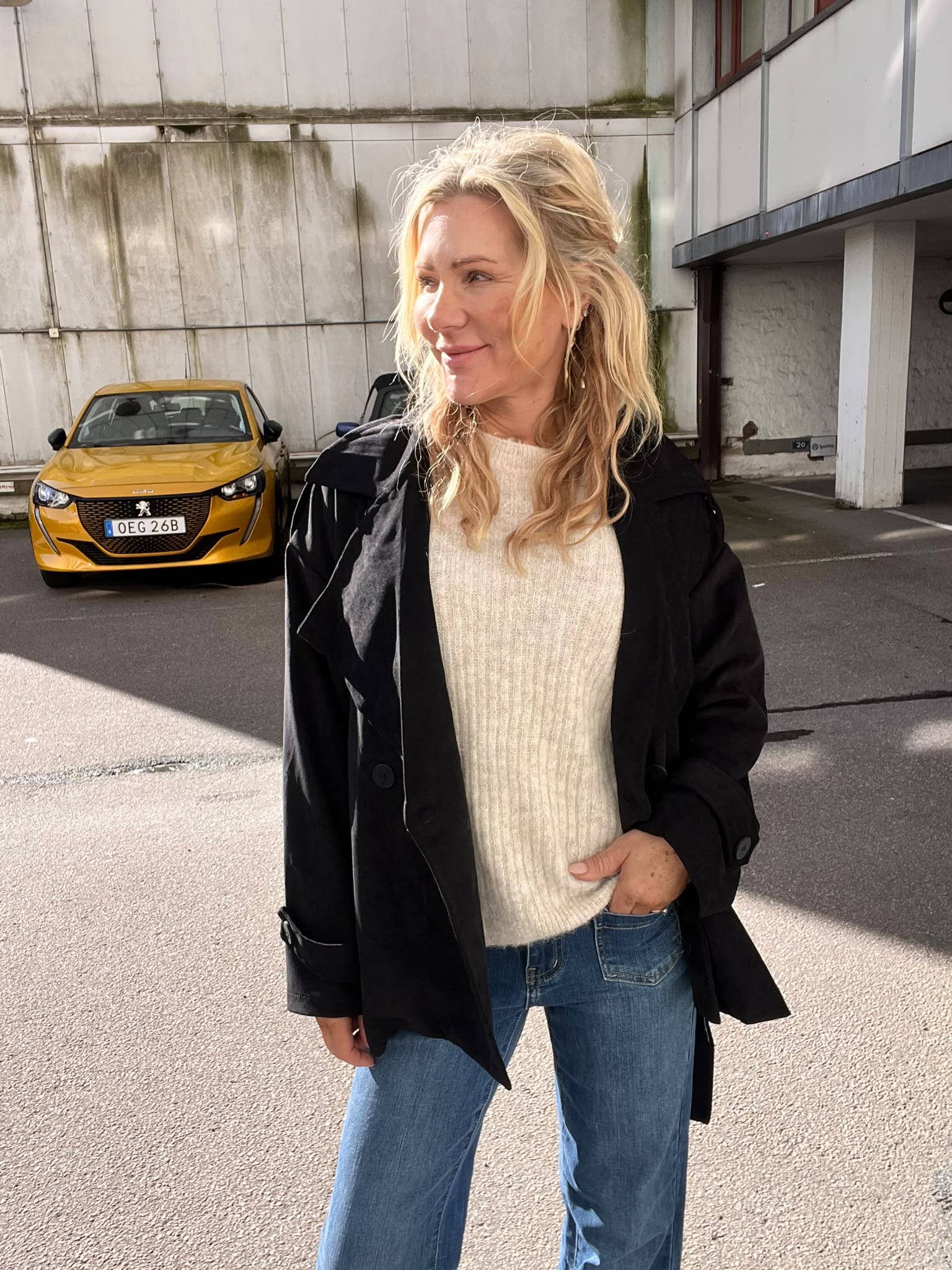 Blond kvinna i svart jacka och ljus stickad tröja står utomhus på en parkeringsplats och tittar åt sidan. Bakom henne syns en gul bil med registreringsskylt “OEG 26B” vid en betongvägg.