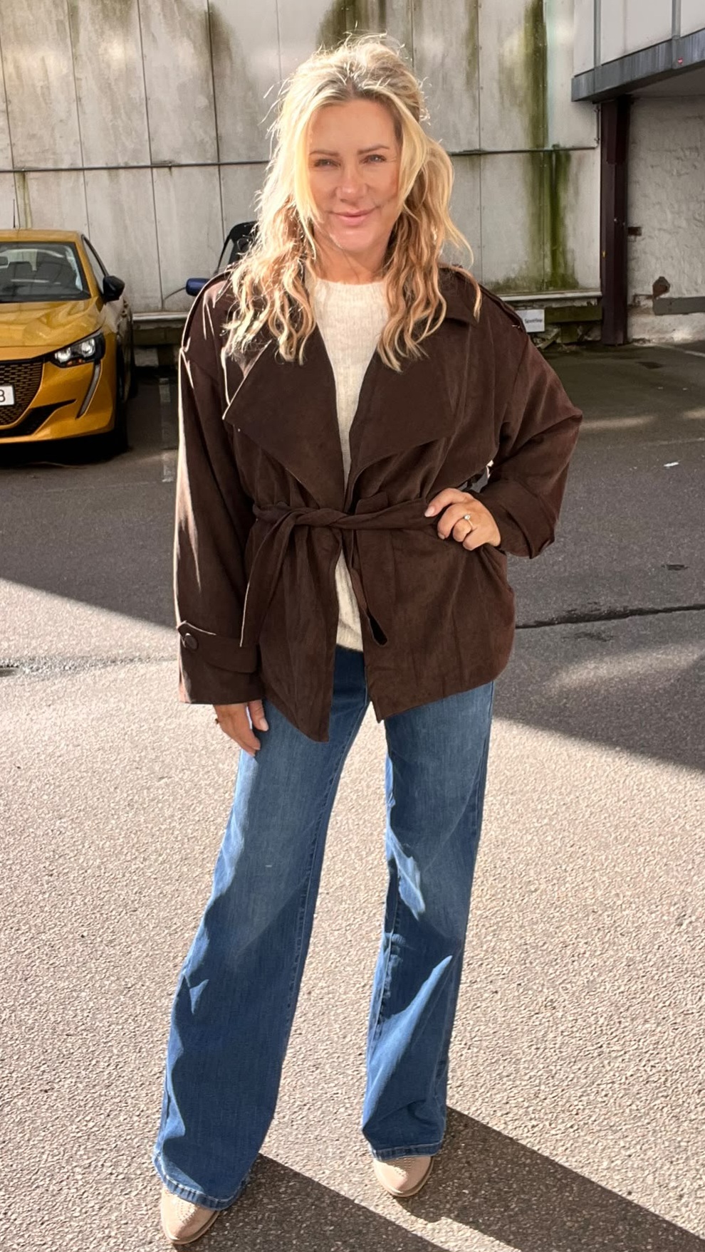 Blond kvinna står och ler mot kameran på en solig parkering, med ena handen på höften. Hon bär brun knuten jacka över ljus tröja, blå utsvängda jeans och ljusa skor; gul bil och betongvägg i bakgrunden.