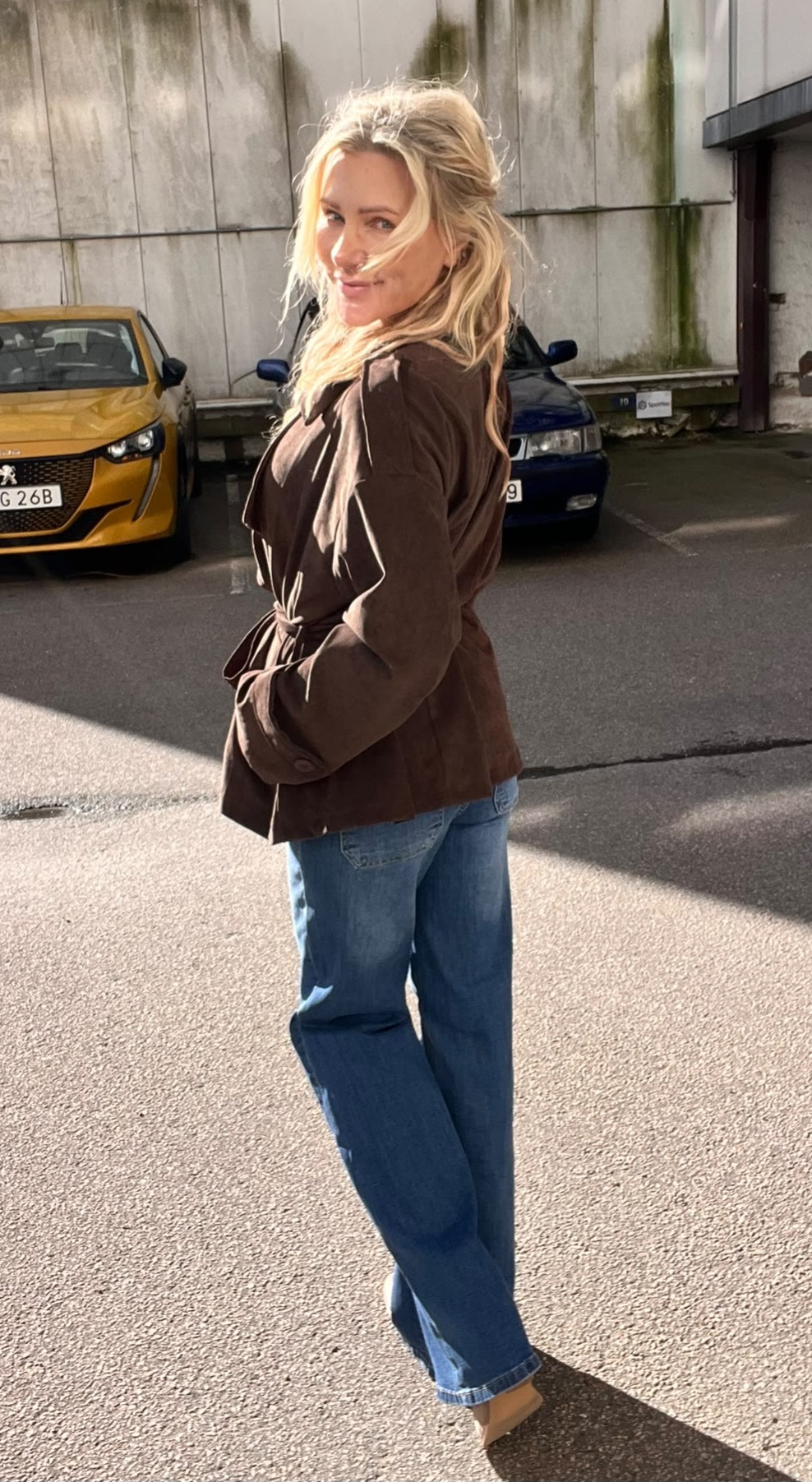 Blond kvinna i brun jacka och blå jeans står utomhus på en parkeringsplats, vänder sig om och ler mot kameran. Solen lyser på henne; bakom syns en betongvägg och två parkerade bilar.