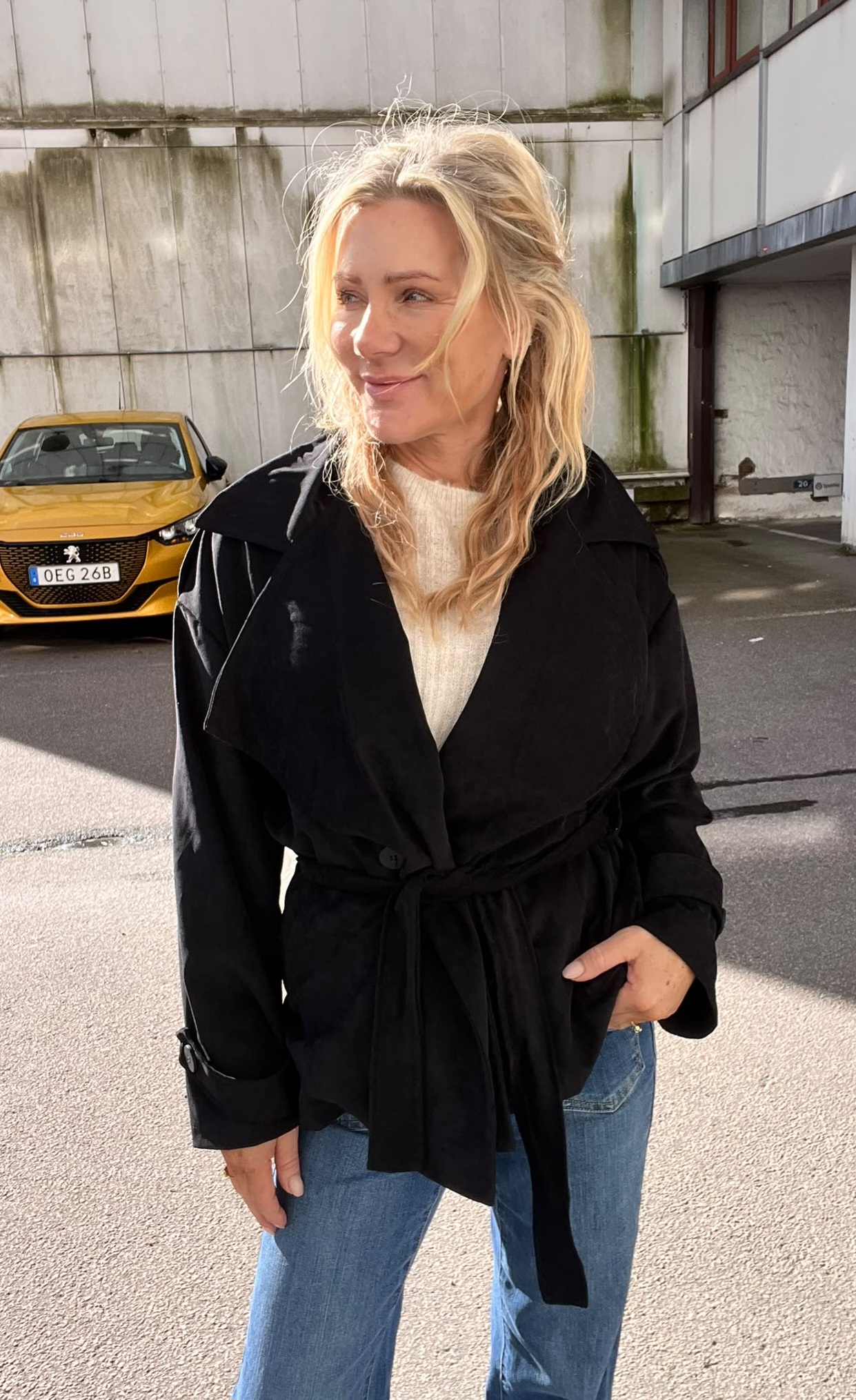 Blond kvinna i svart trenchcoat och jeans står utomhus och tittar åt sidan med ett svagt leende. I bakgrunden syns en gul bil med registreringsskylt “OEG 26B” framför en betongfasad.