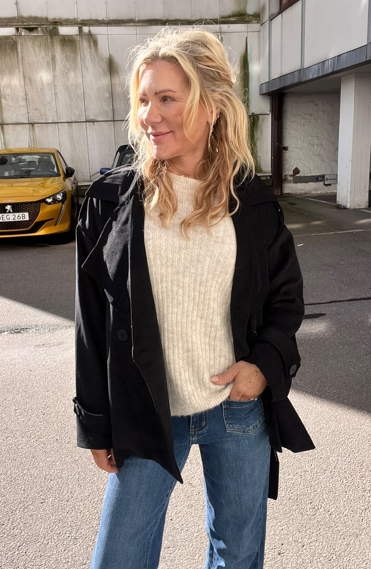 Blond kvinna står utomhus och ler medan hon tittar åt sidan, med handen i jeansfickan. Hon bär svart kappa över beige stickad tröja. I bakgrunden syns en gul bil med registreringsskylt “DEG 26B” och en byggnadsvägg.