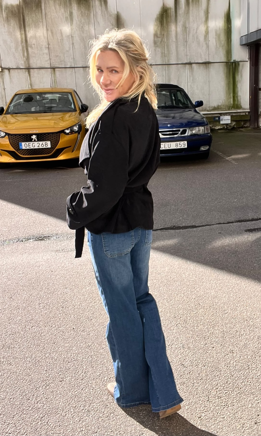 Blond kvinna i svart blus, blå jeans och beige klackar står på en parkeringsplats och vänder sig leende mot kameran. Bakom henne står en gul bil med registreringsskylt “OEG 26B” och en blå bil.