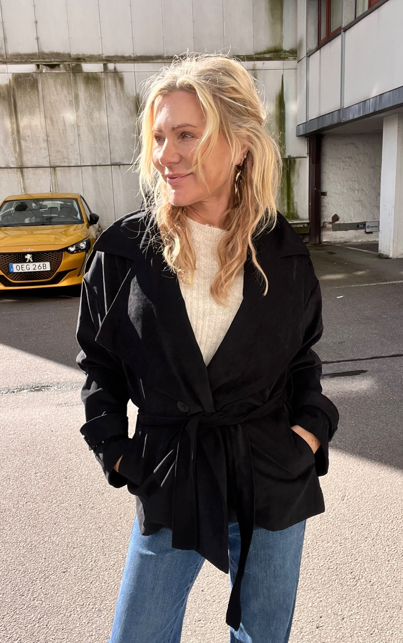 Blond kvinna i svart kappa och ljus stickad tröja står med händerna i fickorna och tittar åt sidan i solsken. I bakgrunden syns en gul bil med registreringsskylt “OEG 26B” vid en betongbyggnad.
