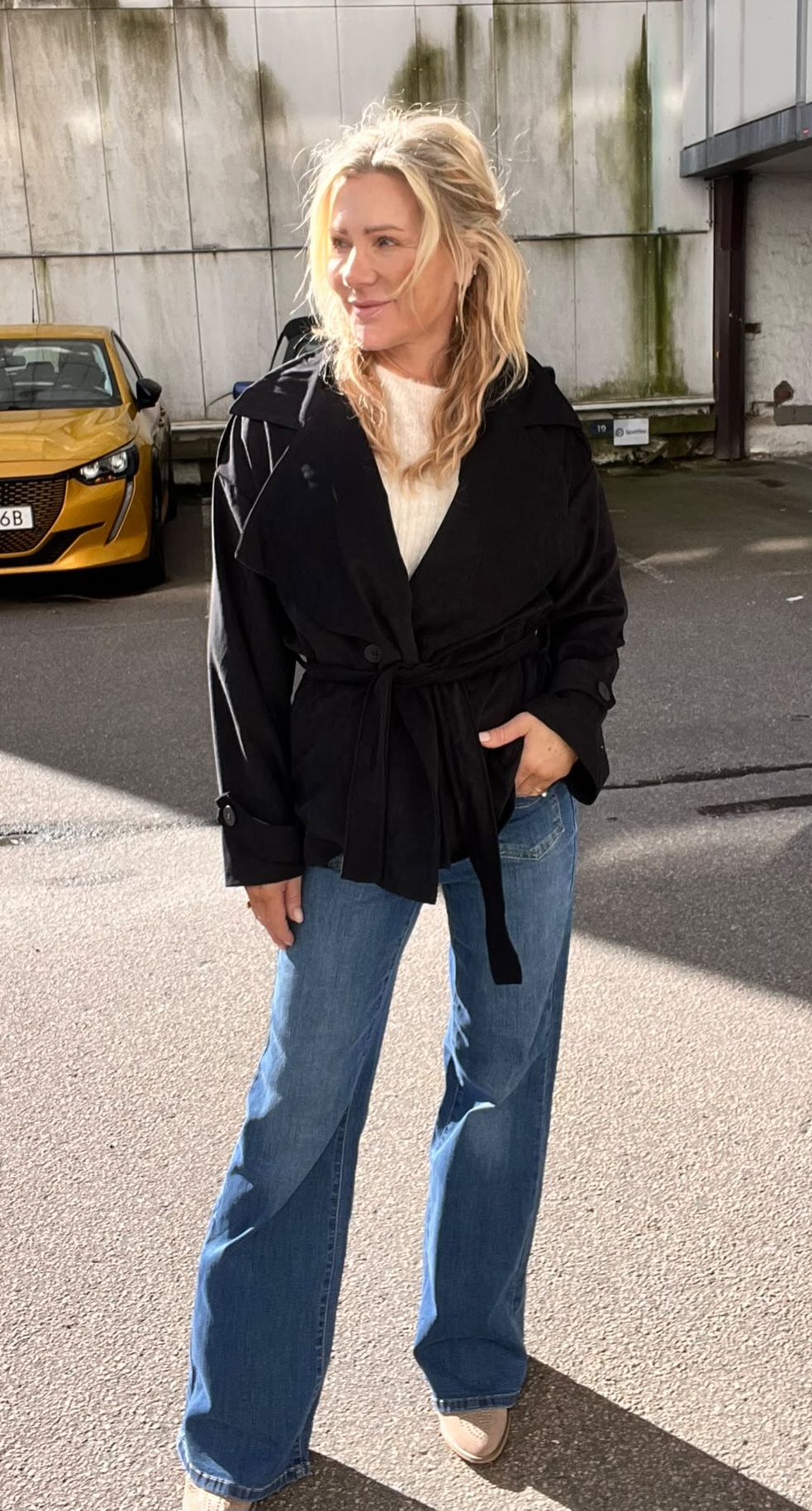 Blond kvinna står utomhus på en asfalterad yta och ler medan hon tittar åt sidan, med ena handen i jeansfickan. Hon bär svart knuten jacka, vit tröja och utsvängda jeans; gul bil och betongvägg bakom.