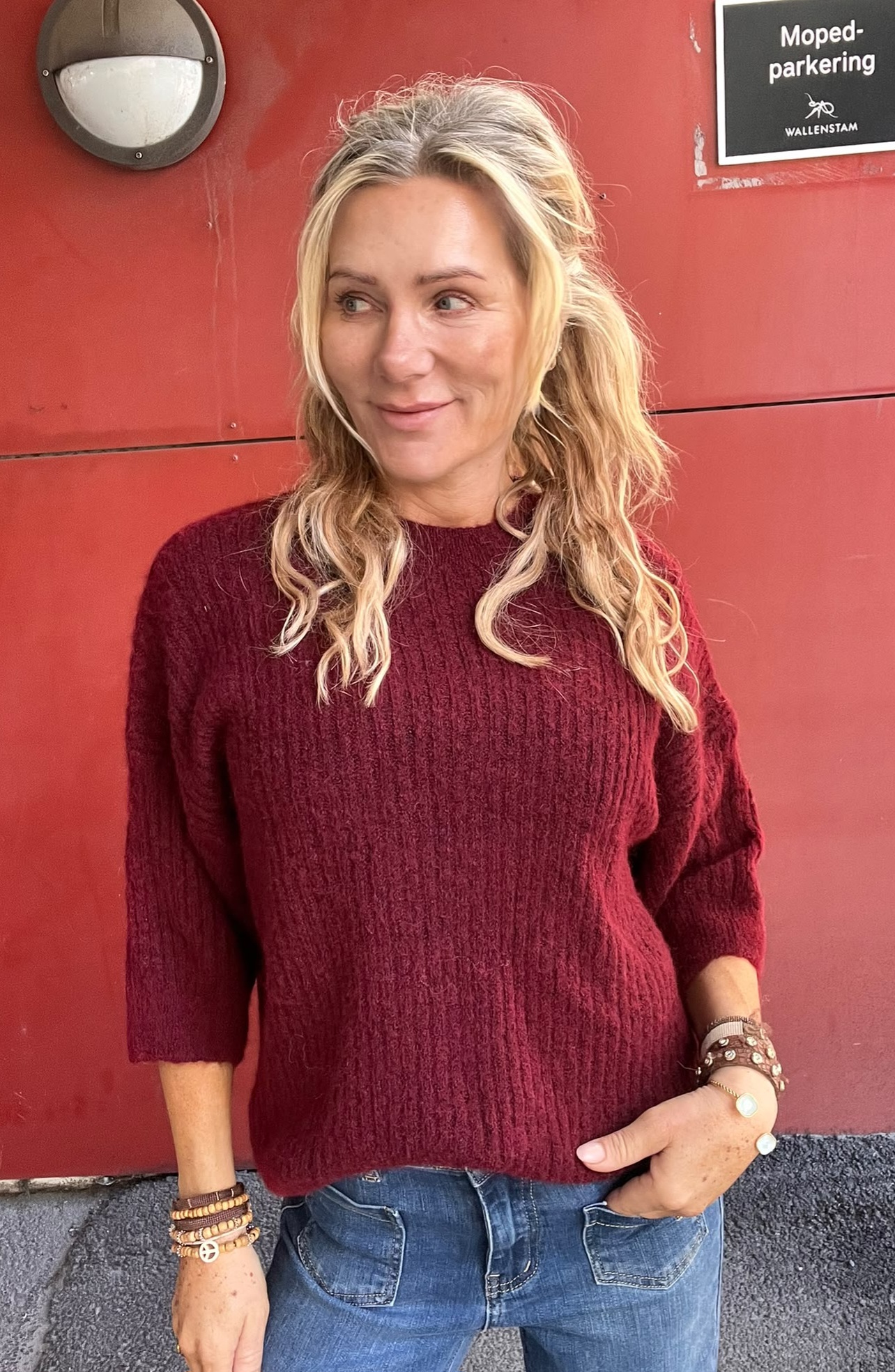 Blond kvinna i vinröd stickad tröja och jeans står avslappnat och tittar åt sidan framför en röd vägg, med handen i fickan. Skylt i bakgrunden: “Moped-parkering” och “WALLENSTAM”.