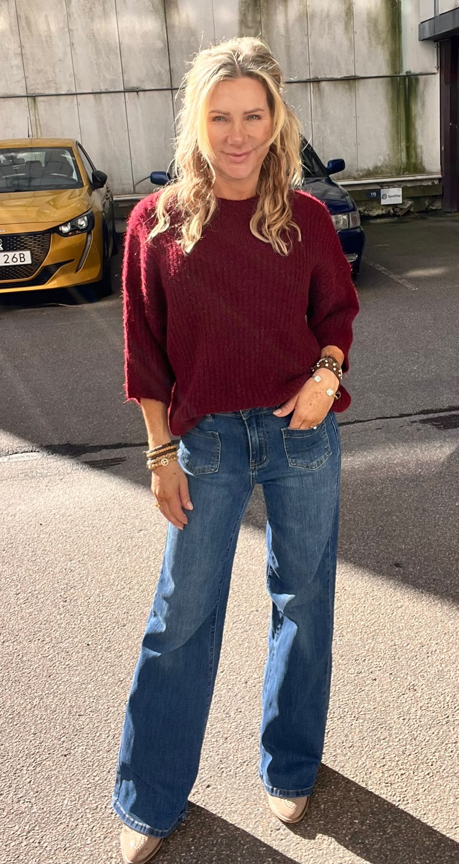 Blond kvinna står och ler mot kameran med en hand i fickan, klädd i vinröd stickad tröja och blå utsvängda jeans med armband. Hon står på en parkeringsplats framför betongvägg och bilar.
