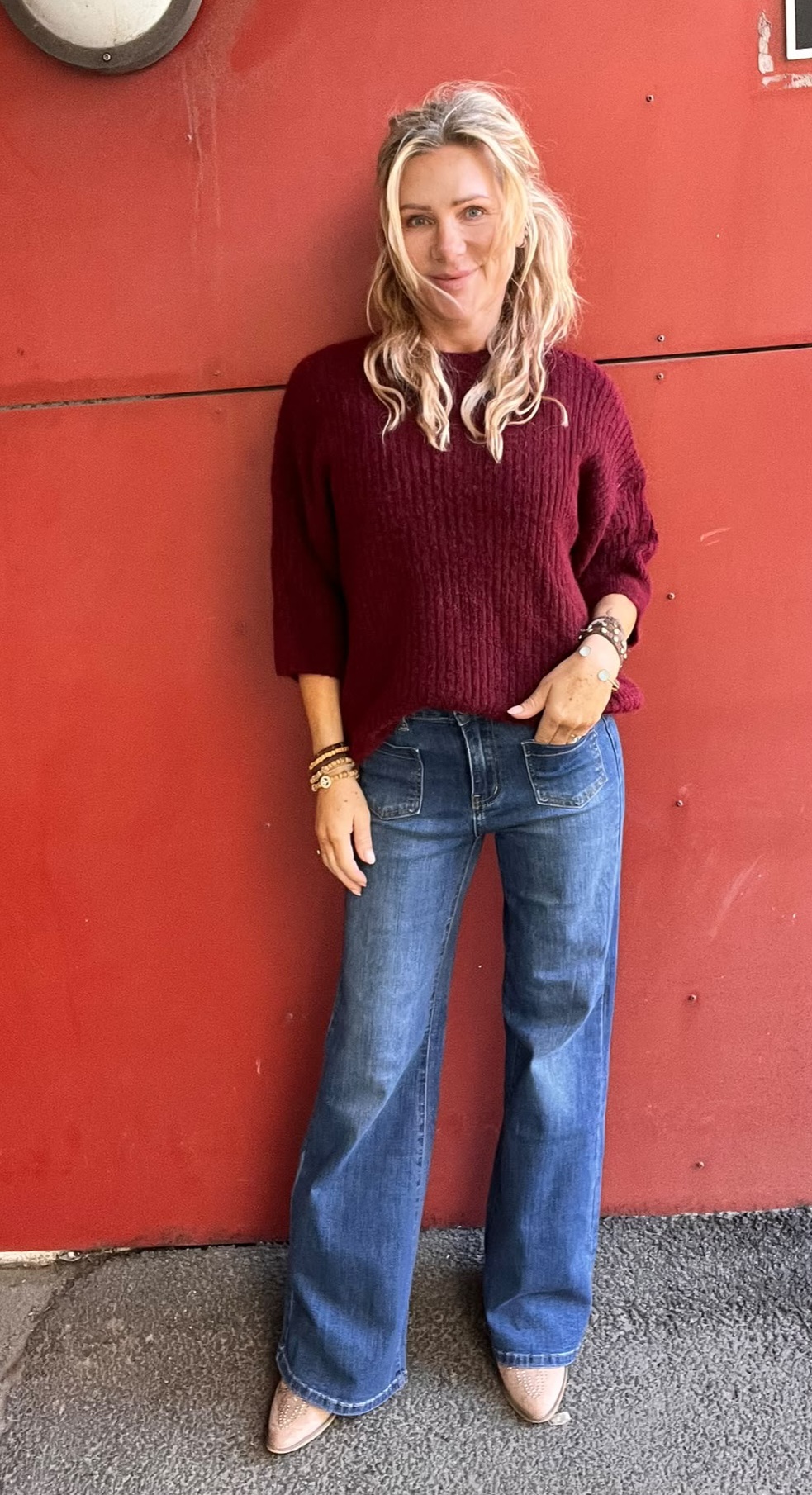 Leende kvinna med långt blont hår står avslappnat mot en röd vägg, med ena handen i jeansfickan. Hon bär vinröd stickad tröja, vida blå jeans, armband och ljusa skor; en rund vägglampa syns ovanför.