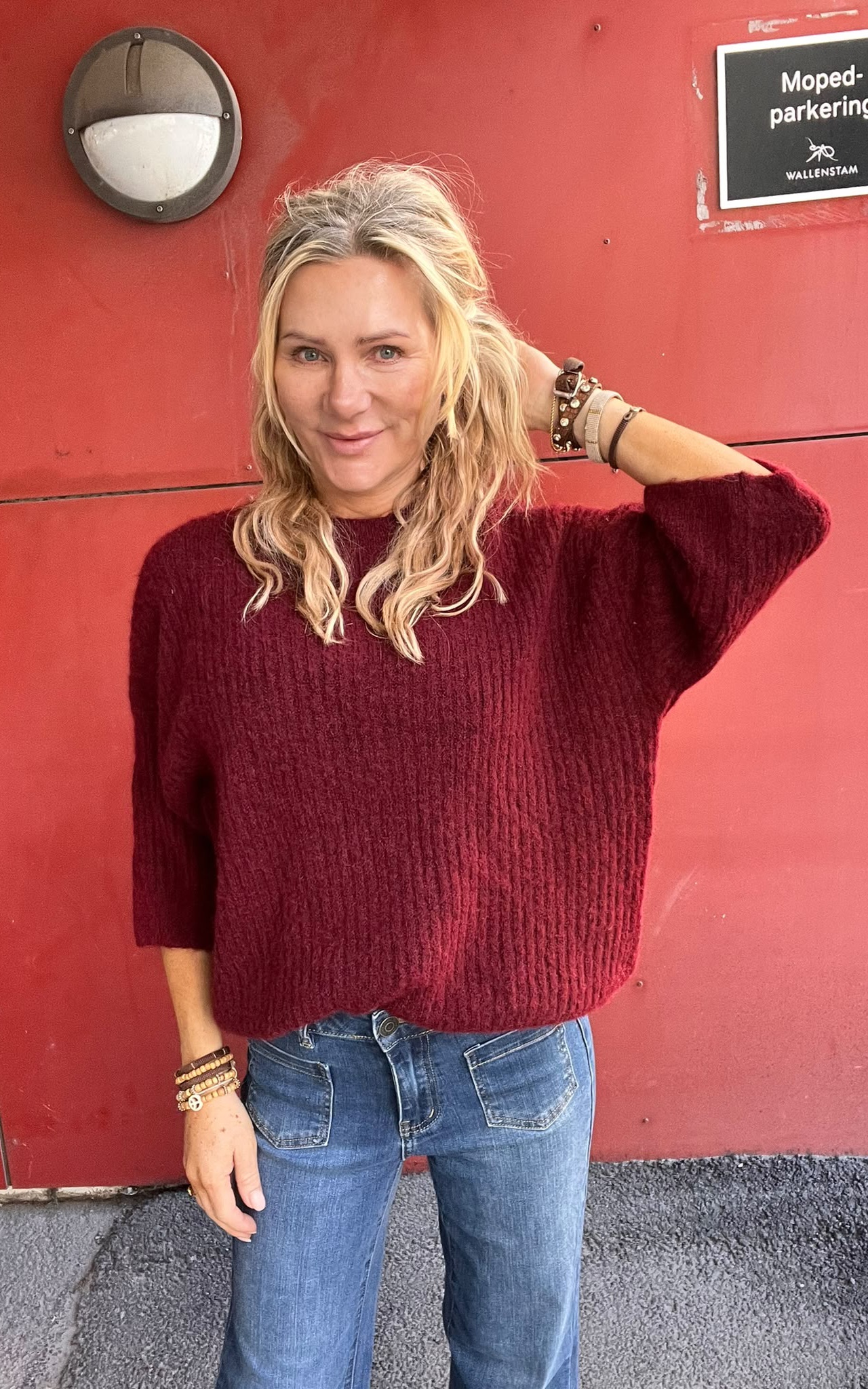 Blond kvinna står och poserar med ena handen bakom huvudet, iförd vinröd stickad tröja och jeans, mot en röd vägg med vägglampa. Skylt med text: “Moped-parkering” och “WALLENSTAM”.
