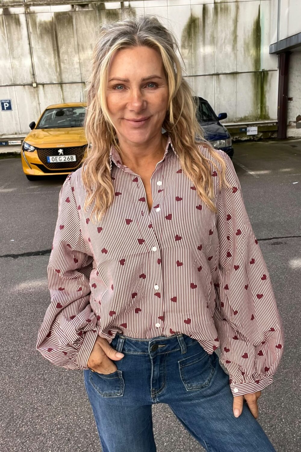 Leende kvinna med långt blont hår står avslappnat med handen i jeansfickan på en parkeringsplats. Hon bär randig skjorta med röda mönster. Bakom henne syns en gul bil med registreringsskylt “OEG 26B” och skylt “PRIVAT P”.