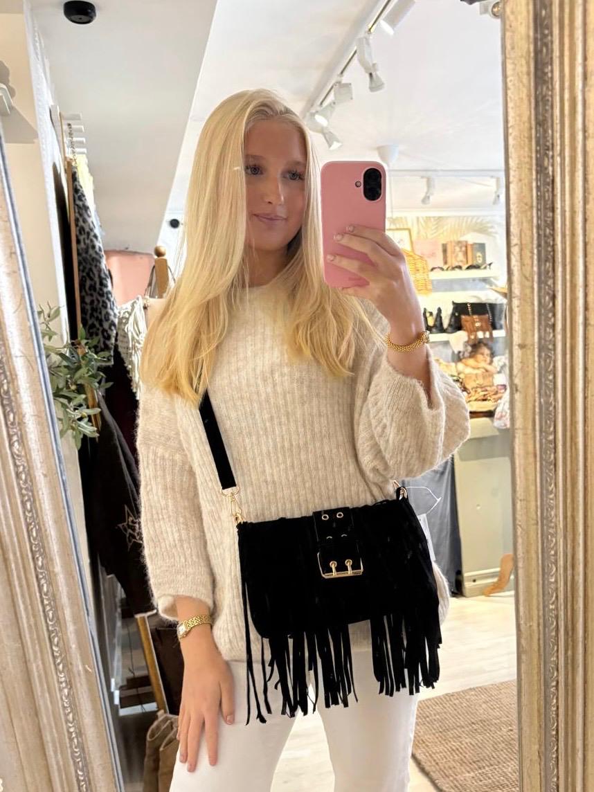 Person med långt blont hår tar en spegelselfie med rosa mobil i en butik, klädd i ljus stickad tröja och vita byxor. En svart fransad axelväska med spänne hänger över kroppen; klädställ och hyllor syns bakom.