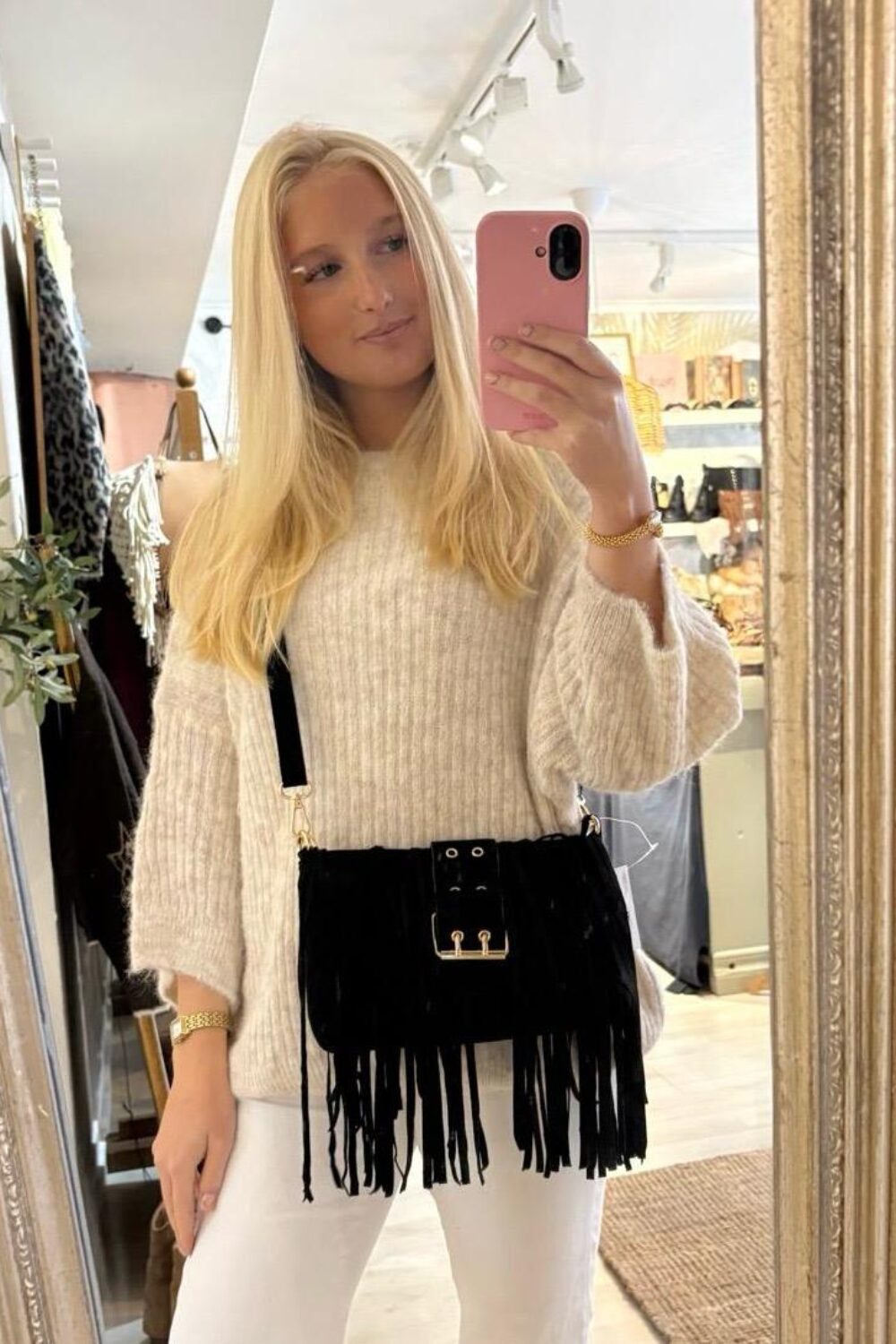 Blond kvinna tar en spegelselfie med rosa mobil i en butik. Hon bär en ljus stickad tröja och vita byxor samt en svart fransad axelväska med gulddetaljer; kläder och väskor syns i bakgrunden.