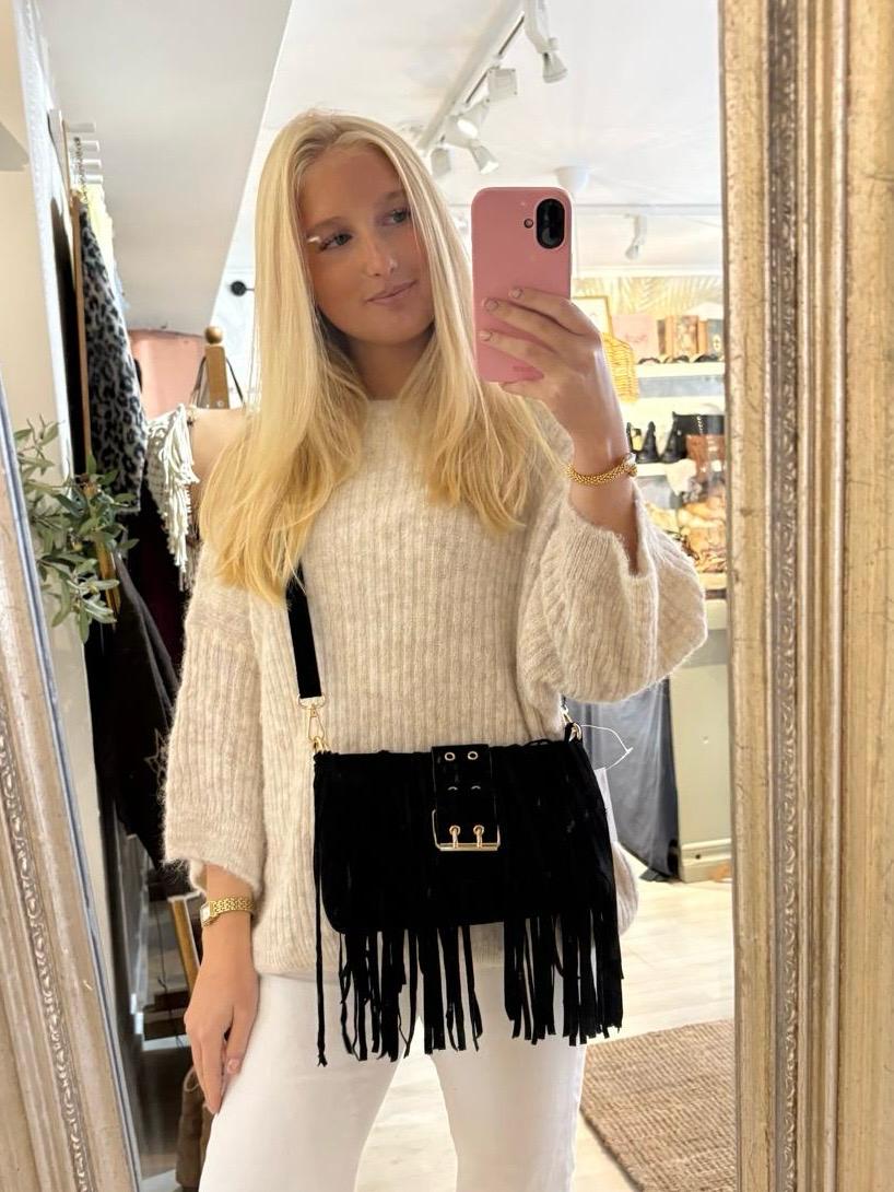 Blond kvinna tar en spegelselfie med rosa mobil i en butik. Hon bär en ljus stickad tröja och vita byxor samt en svart fransad axelväska med gulddetaljer; kläder och väskor syns i bakgrunden.