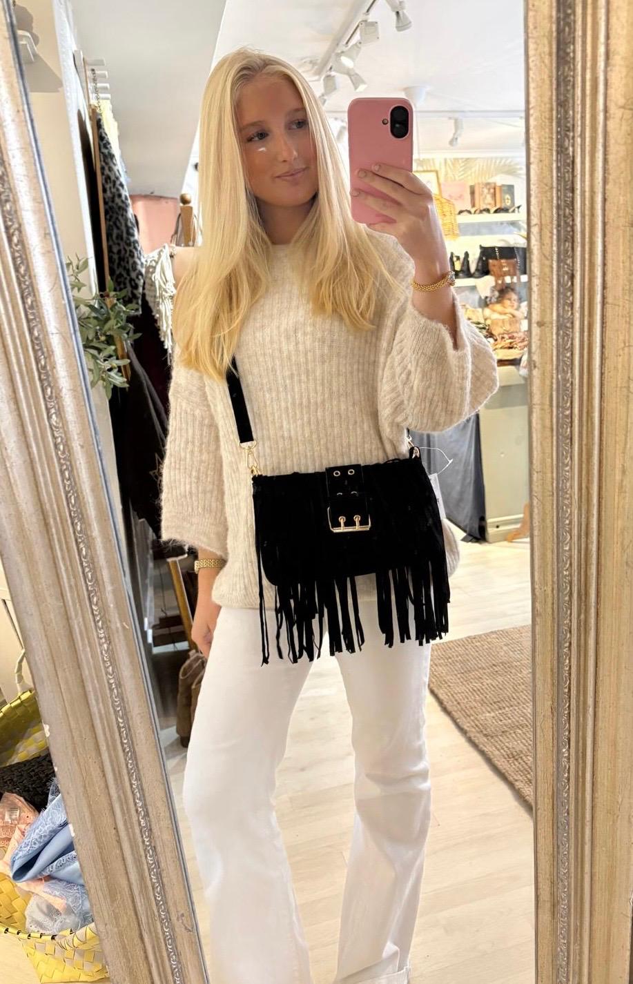 Blond person tar en spegelselfie med rosa mobil i en butik, klädd i beige stickad tröja och vita byxor. En svart axelväska med fransar hänger över kroppen, hyllor med skor syns i bakgrunden.