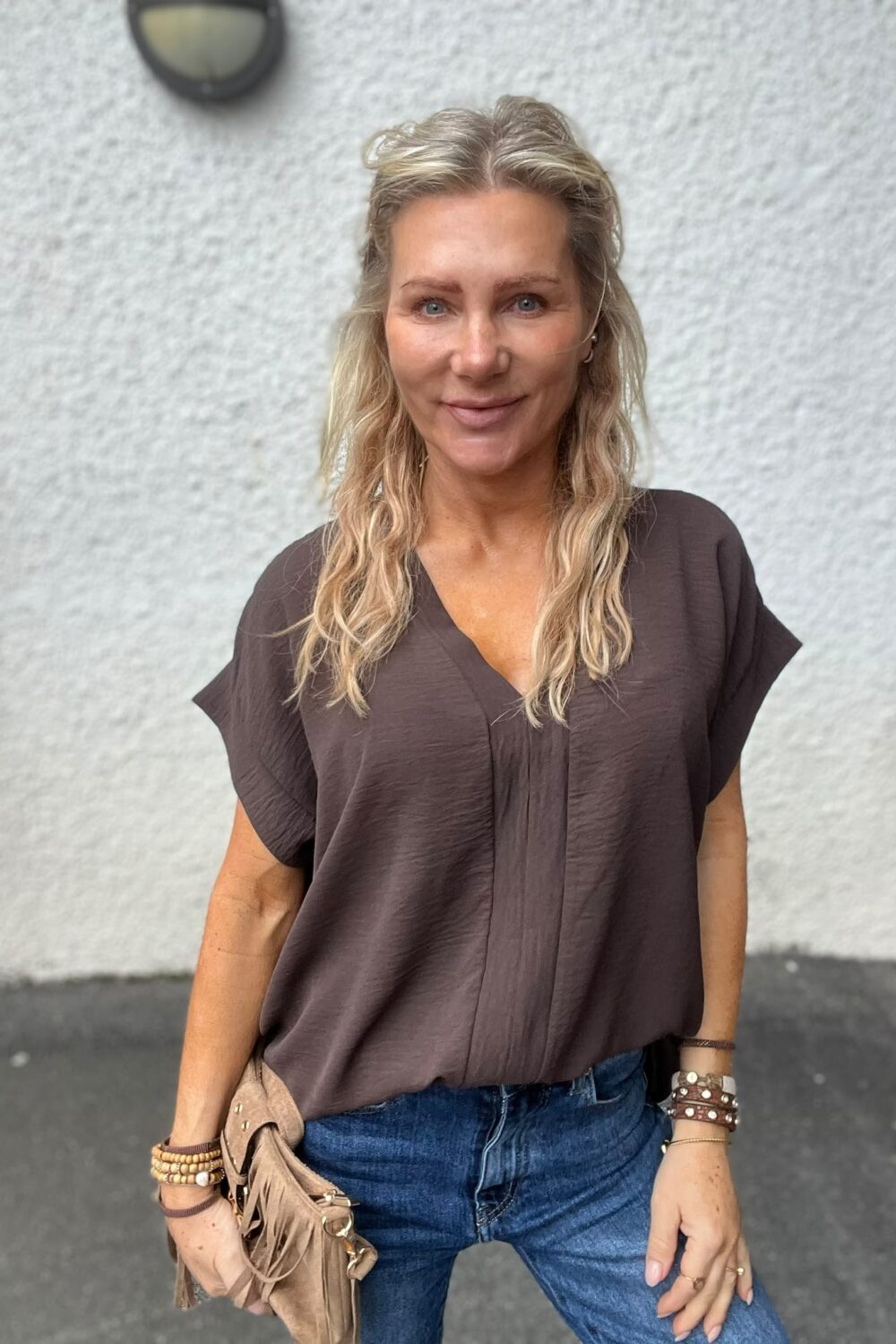 Leende kvinna med långt blont hår står avslappnat utomhus framför en ljus, grovputsad vägg, med ena handen i jeansfickan. Hon bär brun blus, armband och en beige fransad väska.