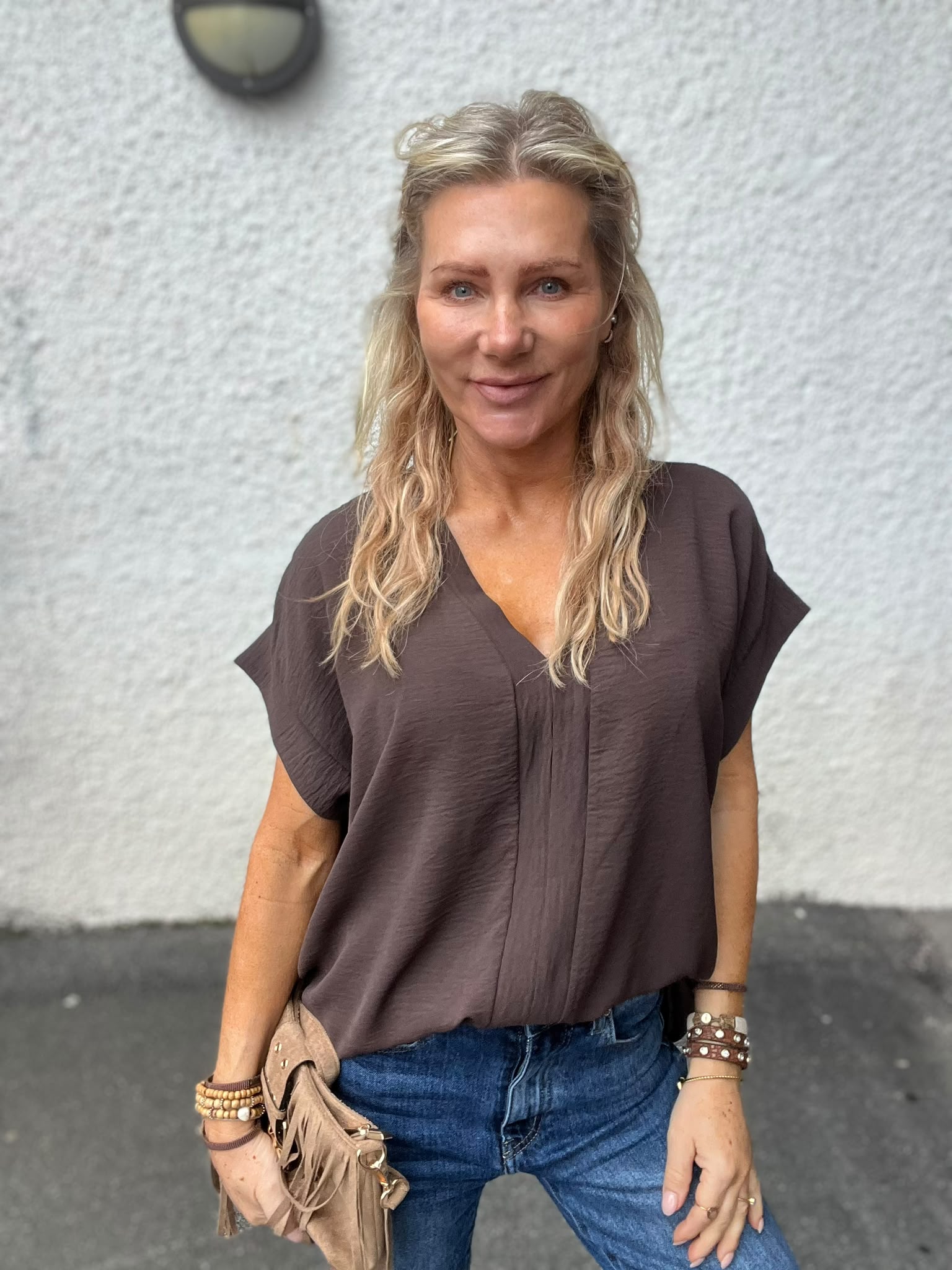 Blond kvinna står utomhus och ler mot kameran, med händerna i jeansfickorna. Hon bär brun, lös blus, flera armband och en beige fransväska, framför en ljus putsad vägg med rund vägglampa.