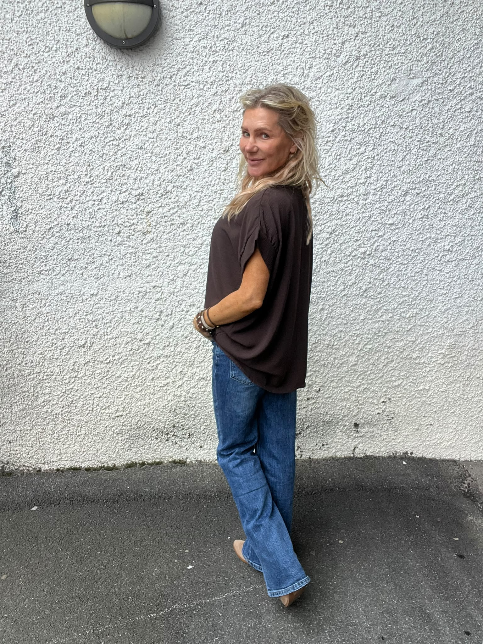 Blond kvinna i mörk, löst sittande topp och blå jeans går längs en grov, vit yttervägg och vänder sig om mot kameran med ett leende; vägglampa syns ovanför på asfaltmark.