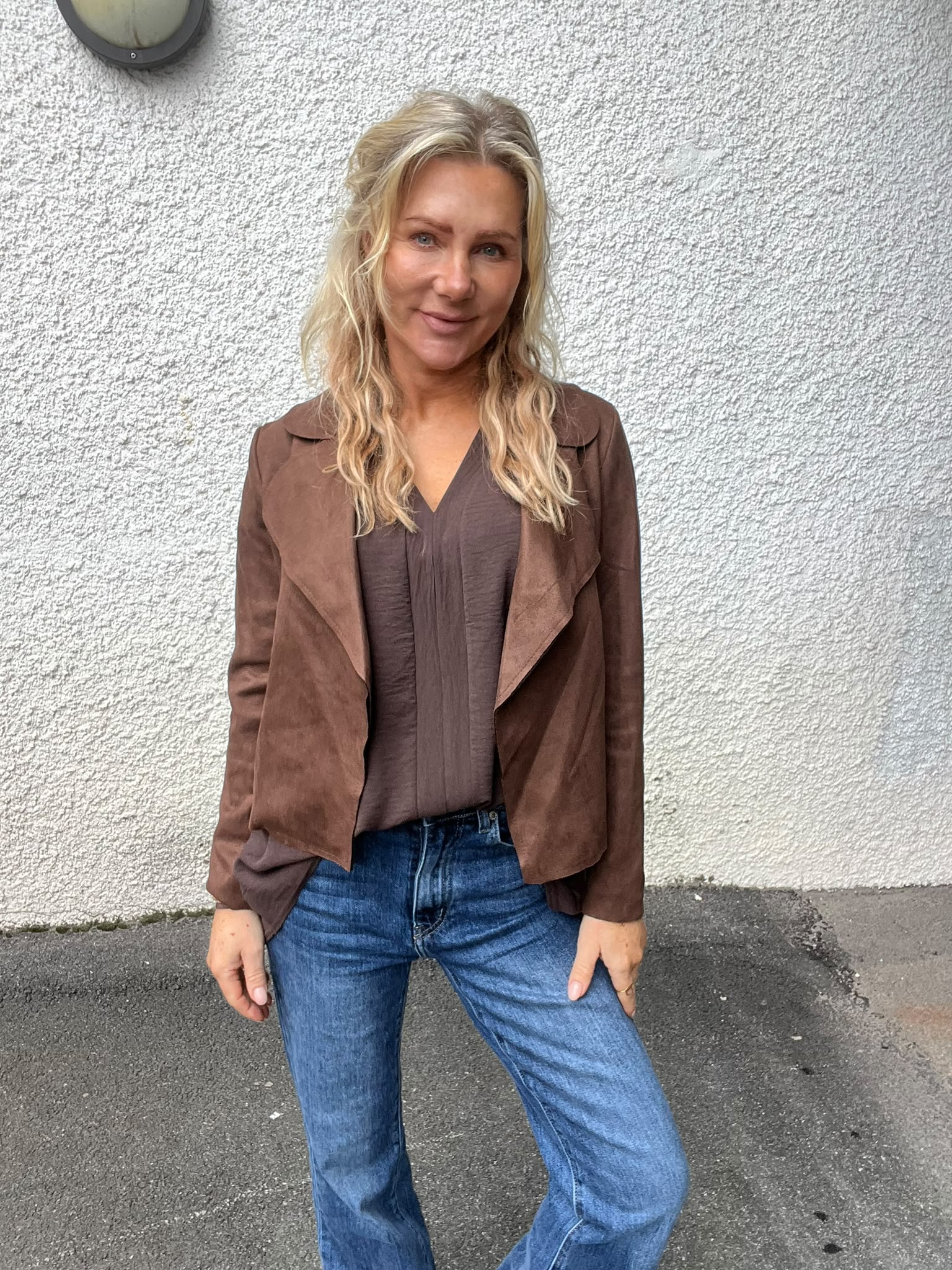 Kvinna med långt blont hår står avslappnat och ler mot kameran. Hon bär brun mockajacka, mörk blus och blå jeans, och står utomhus framför en vit, grovputsad vägg med en lamparmatur.