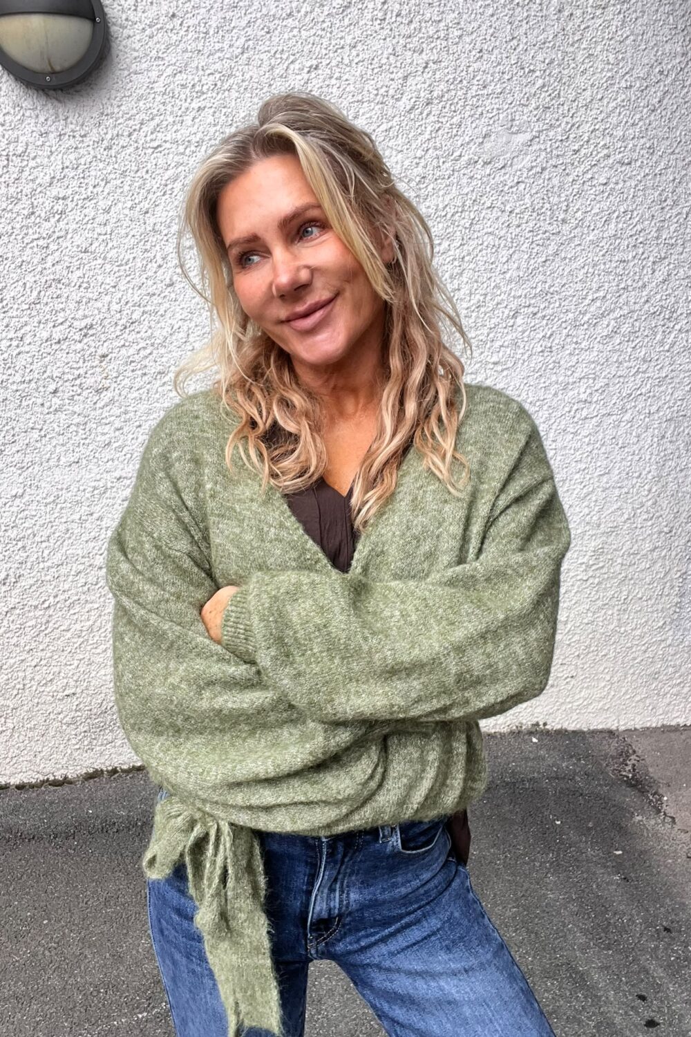 Blond kvinna står med armarna i kors och ler lätt medan hon tittar åt sidan. Hon bär en grön stickad kofta och jeans, framför en ljus, grovputsad yttervägg med en rund vägglampa ovanför.