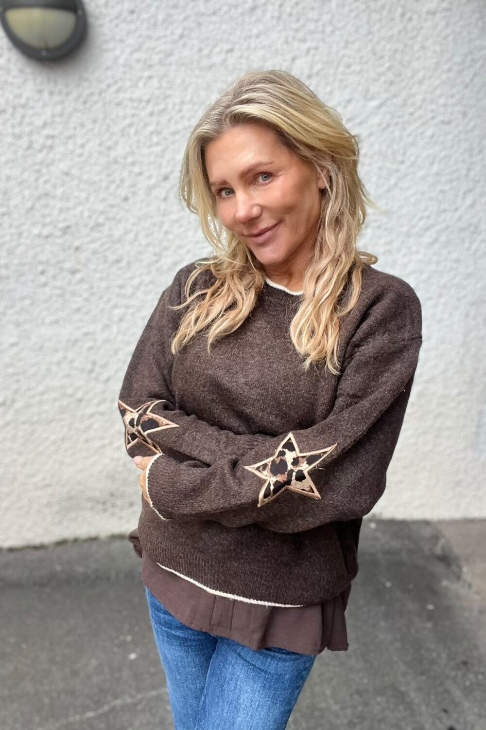 Blond kvinna står med armarna i kors och ler svagt mot kameran. Hon bär brun stickad tröja med stjärnpatchar på armbågarna och jeans, framför en ljusgrå vägg utomhus.