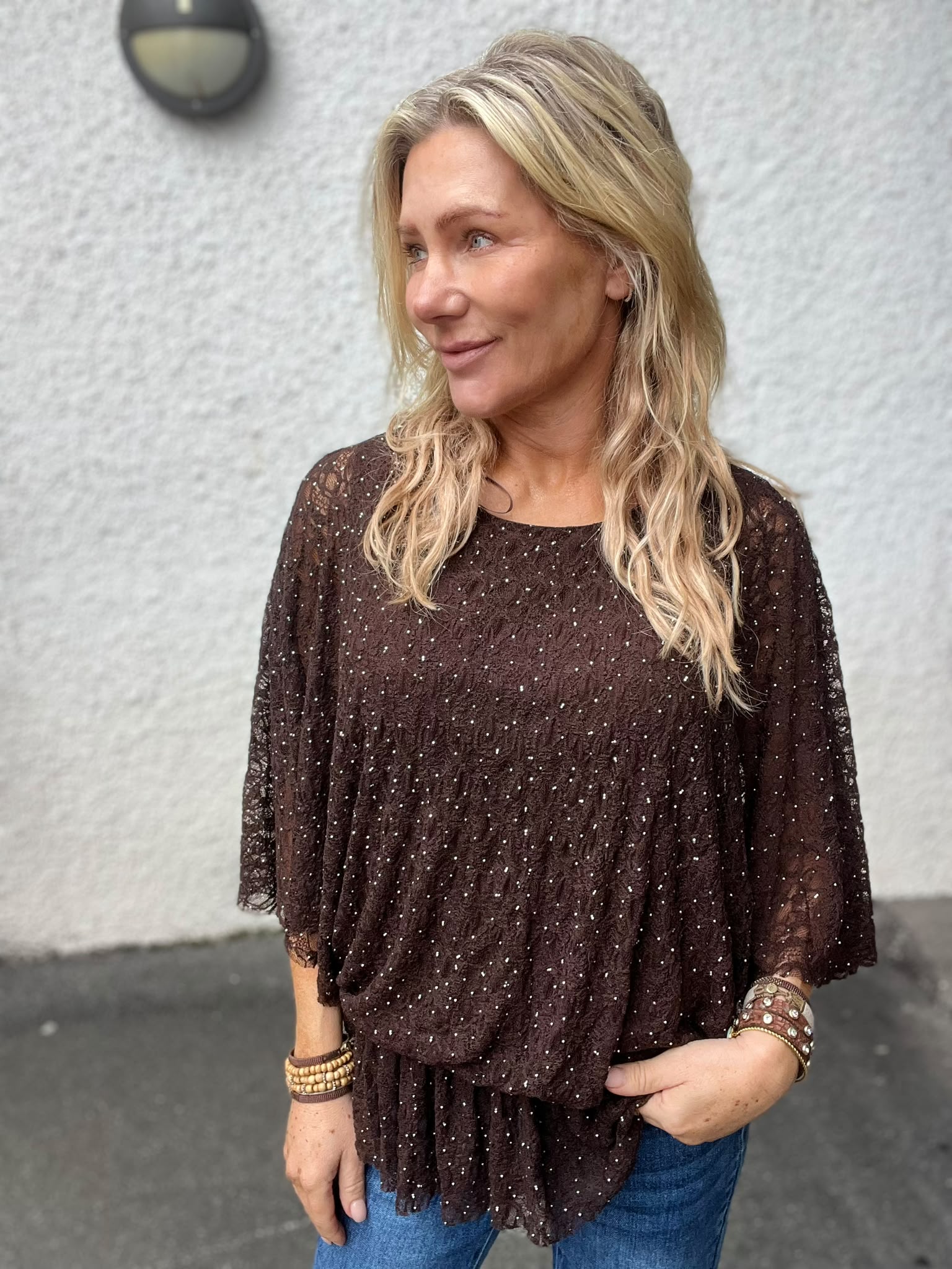 Blond kvinna står utomhus mot en ljus, grovputsad vägg och tittar åt sidan med ett svagt leende. Hon bär en mörkblond spetsblus med små glittriga prickar, jeans och flera armband.