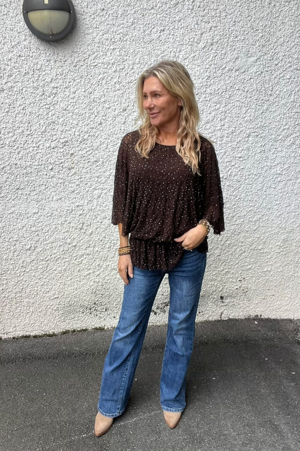 Kvinna med långt blont hår står avslappnat och tittar åt sidan, med ena handen i jeansfickan. Hon bär mörk, prickig blus och beige skor, framför en grov vit vägg med rund vägglampa.