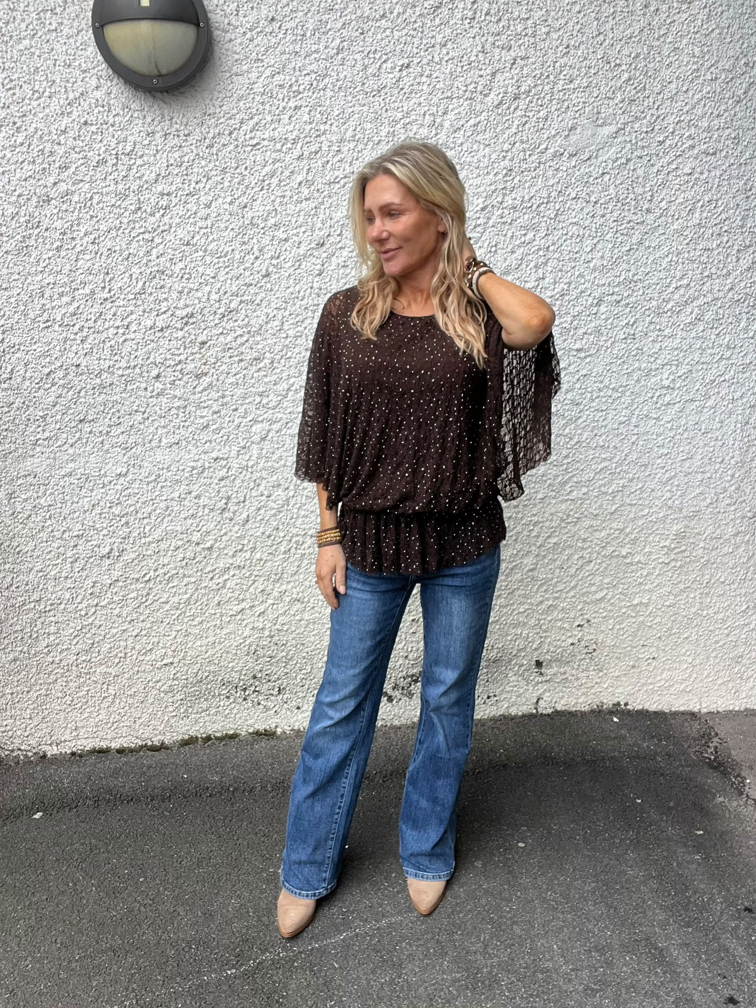 Blond kvinna står avslappnat mot en vit, grovputsad vägg och ser åt sidan med ena handen i håret. Hon bär mörk glittrig blus med vida ärmar, blå jeans och beige skor; vägglampa ovanför.