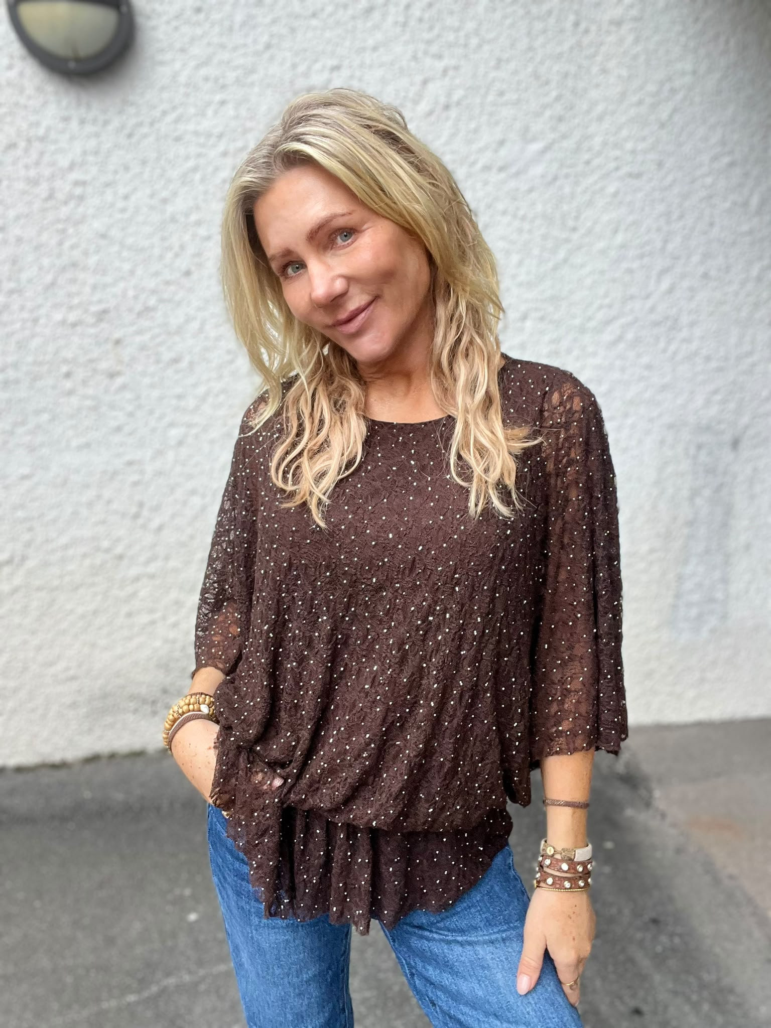 Blond kvinna ler och lutar huvudet lätt åt sidan, med ena handen i jeansfickan. Hon bär en brun, glittrig blus med vida ärmar och flera armband, framför en ljusgrå vägg utomhus.