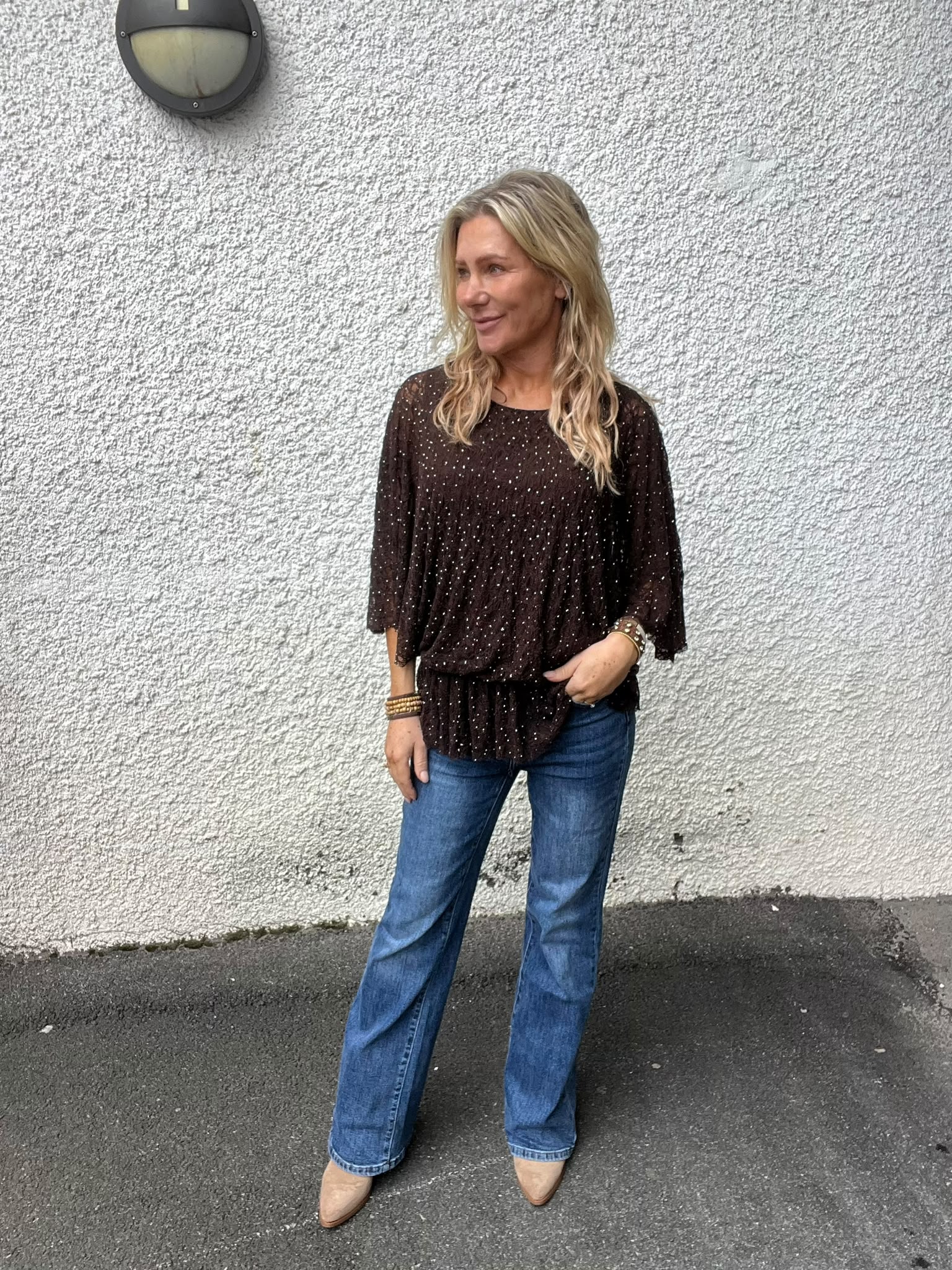 Kvinna med långt blont hår står avslappnat och tittar åt sidan, med ena handen i jeansfickan. Hon bär mörk, prickig blus och beige skor, framför en grov vit vägg med rund vägglampa.