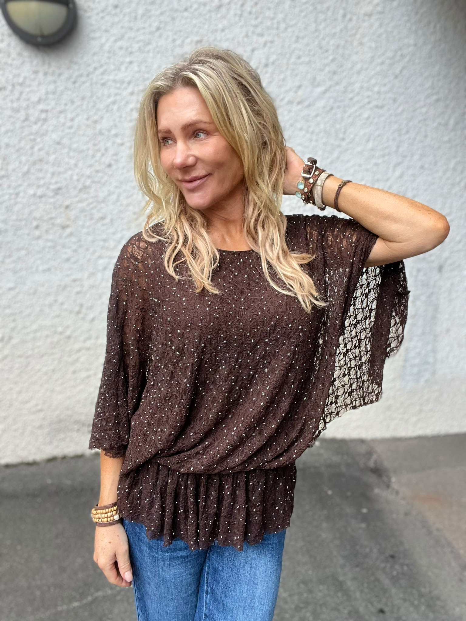 Blond kvinna står utomhus mot en ljus vägg och ler svagt medan hon håller ena handen bakom huvudet. Hon bär brun, glittrig spetsblus med vida ärmar, blå jeans och flera armband.