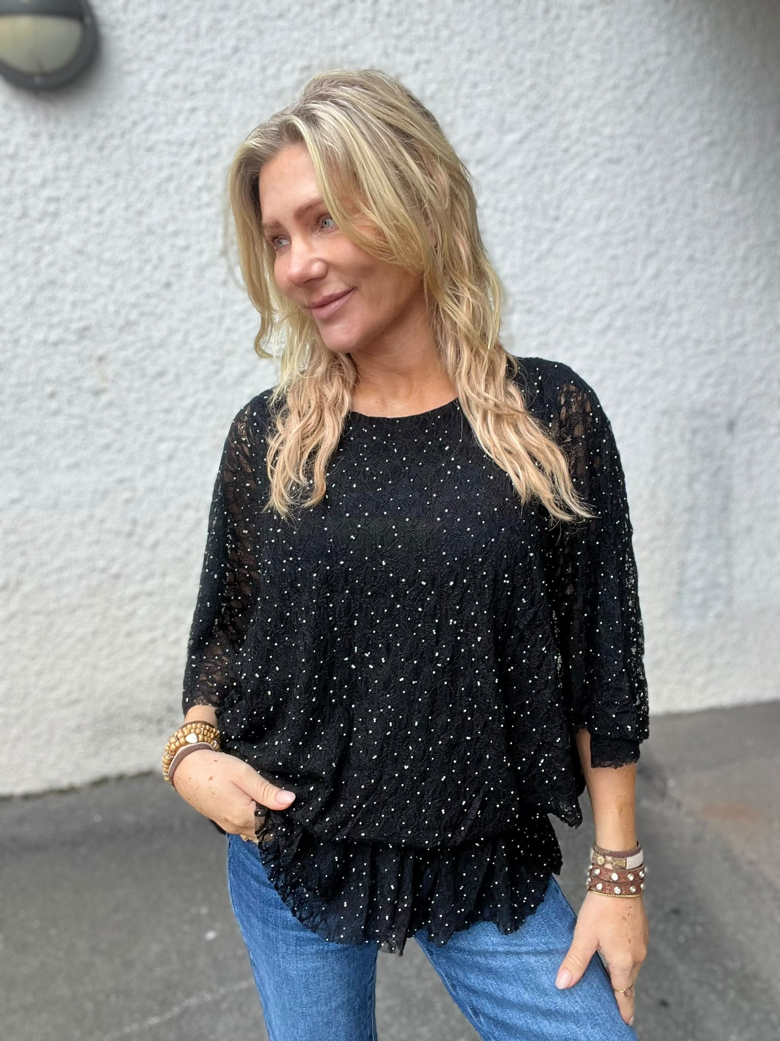 Blond kvinna står utomhus mot en ljus, grov vägg och ler medan hon tittar åt sidan. Hon har en svart, glittrig topp med trekvartsärm, blå jeans och flera armband, med ena handen i fickan.