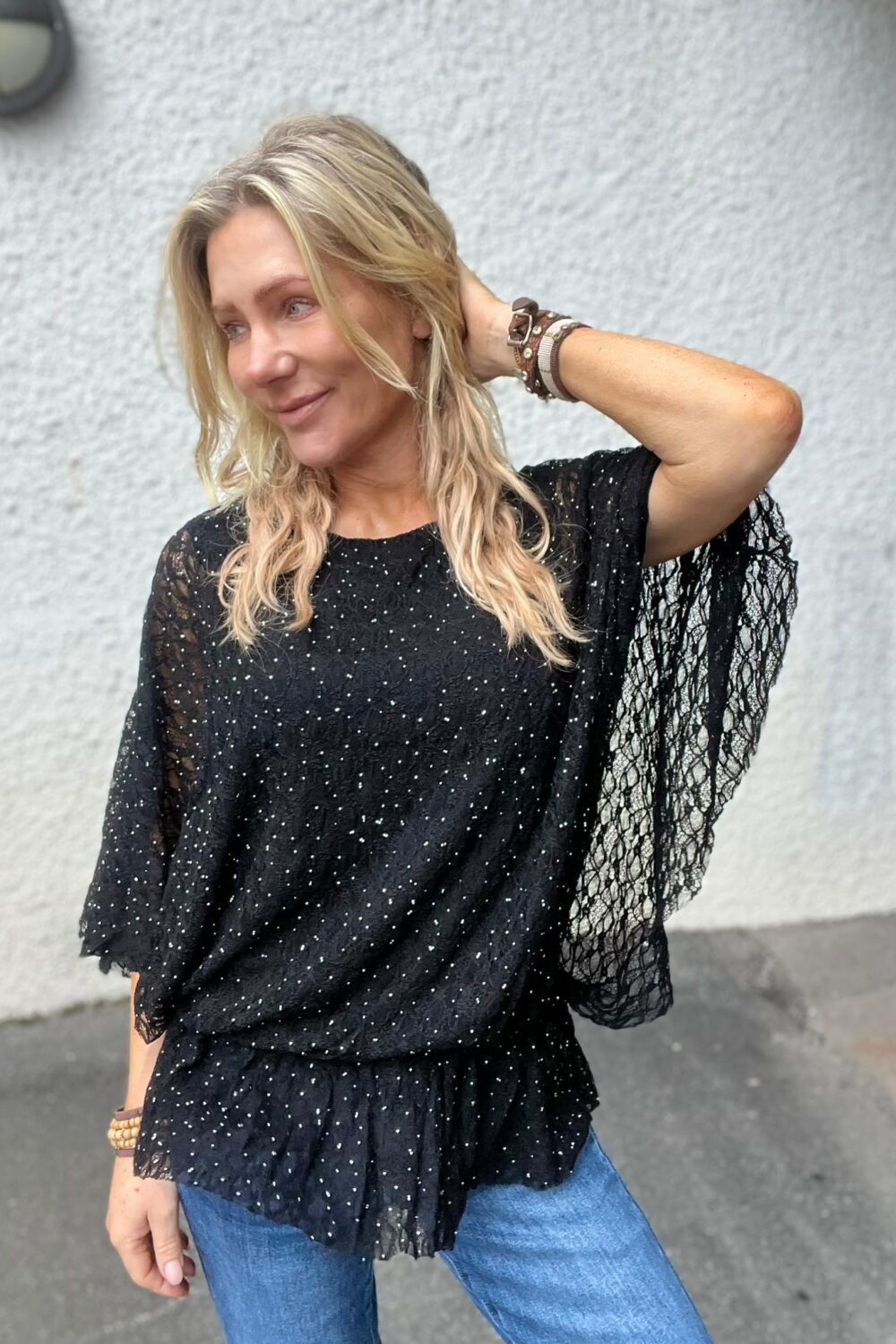 Blond kvinna poserar utomhus med ena handen bakom huvudet och ler åt sidan. Hon bär en svart, glittrig blus med spetsärmar och armband, framför en ljus putsad vägg.