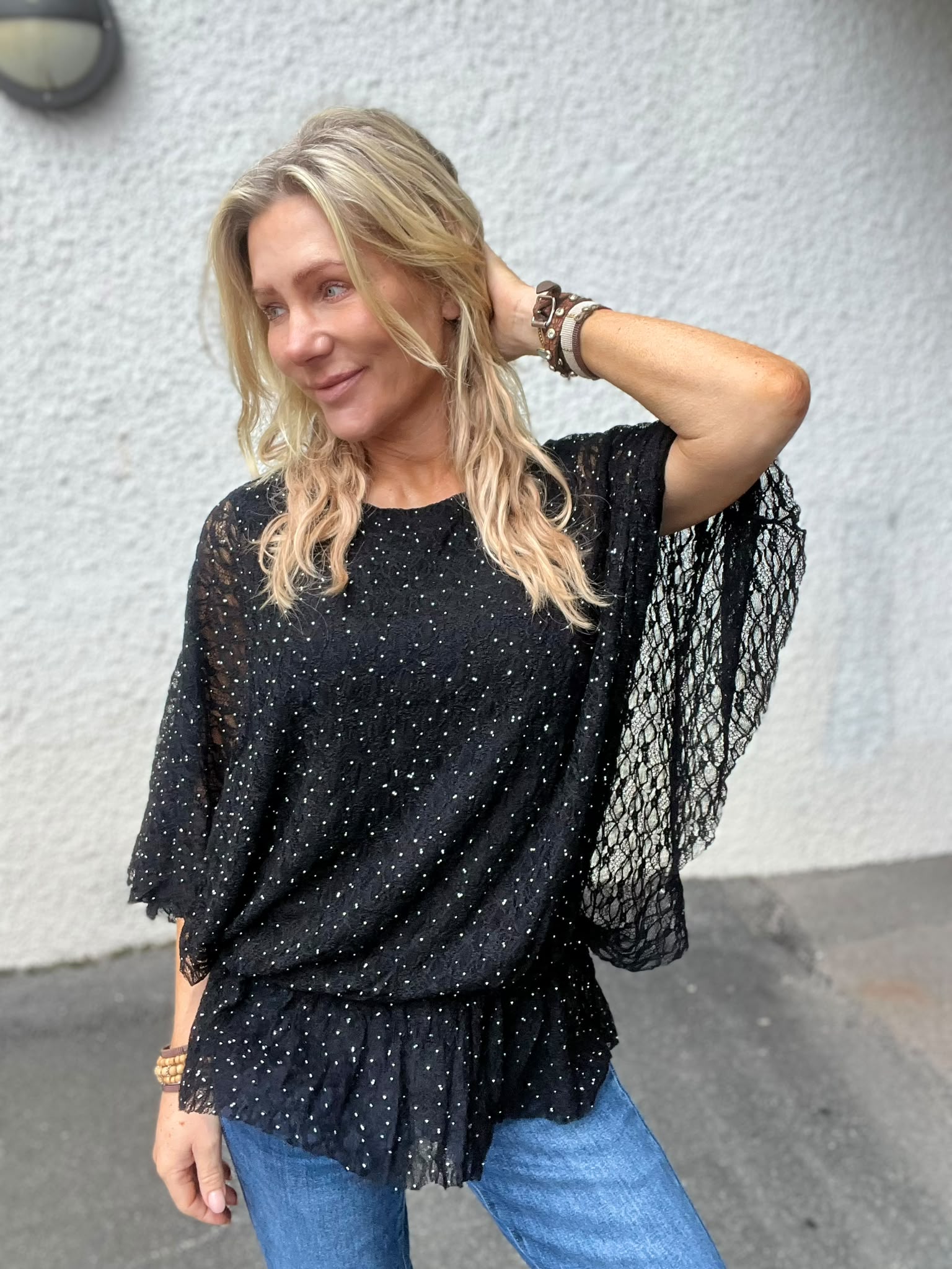 Blond kvinna poserar utomhus med ena handen bakom huvudet och ler åt sidan. Hon bär en svart, glittrig blus med spetsärmar och armband, framför en ljus putsad vägg.
