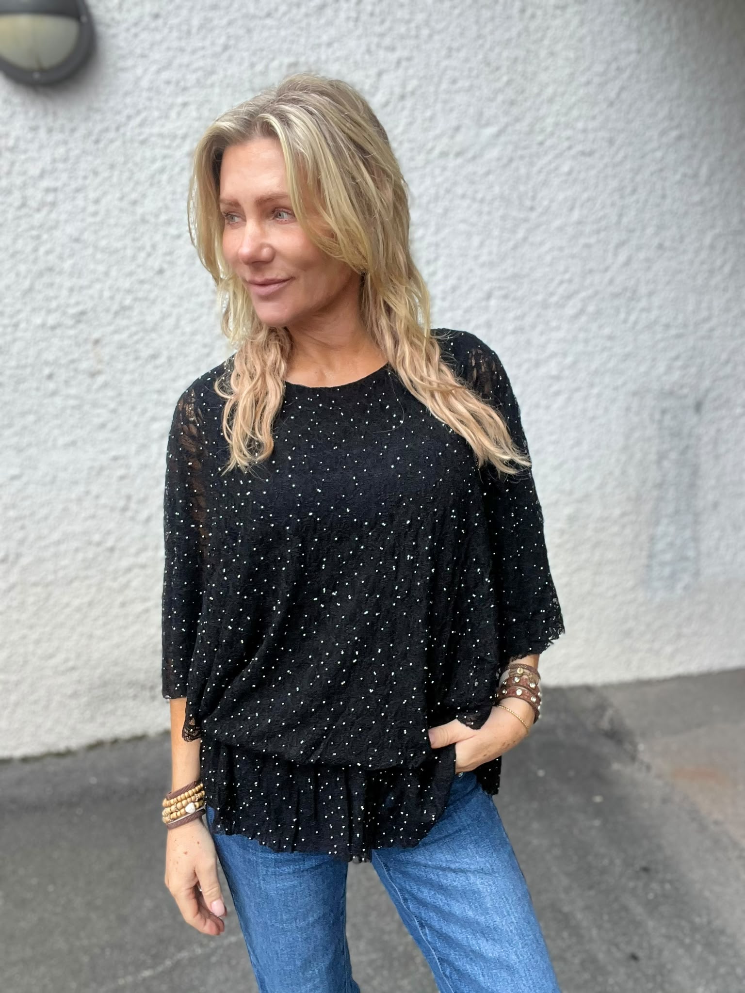 Blond kvinna står avslappnat med ena handen i jeansfickan och tittar åt vänster, klädd i svart glittrig blus och armband. Hon står utomhus framför en ljus, grovputsad vägg och asfalt.
